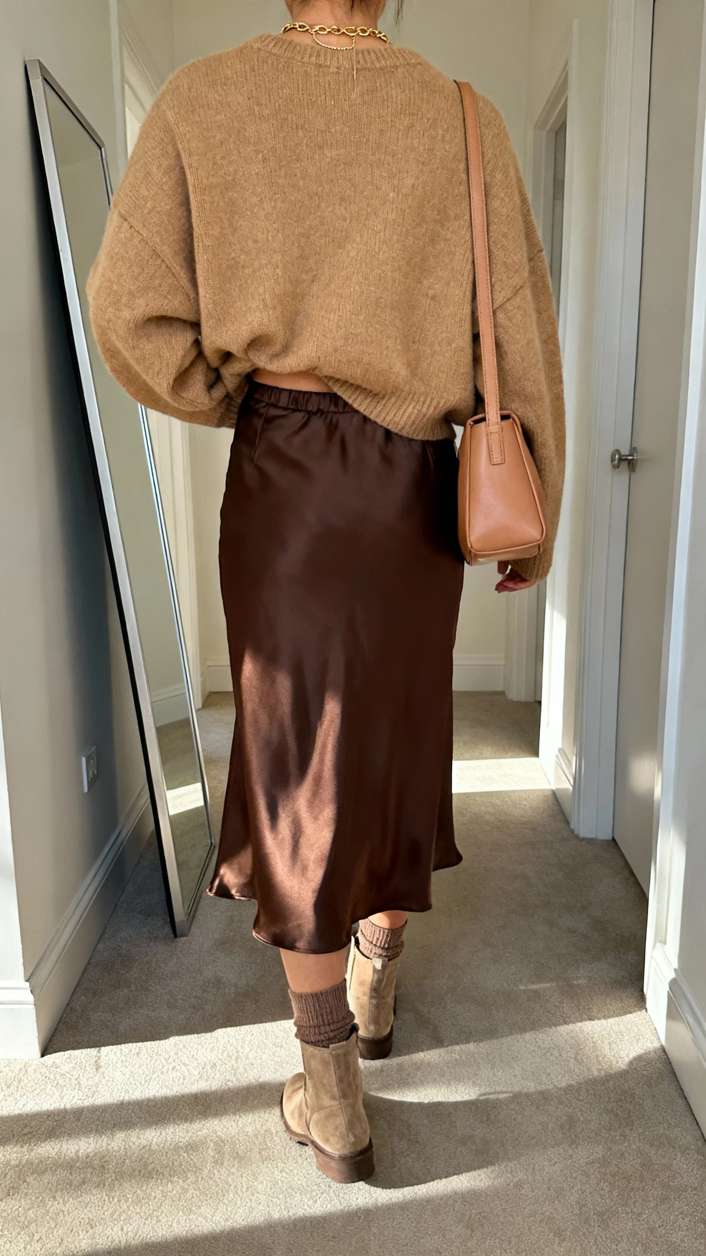iPhone back view of a woman wearing an oversized camel sweater half-tucked into a satin midi skirt in chocolate brown, ankle sock boots, layered dainty gold necklace, and a soft leather shoulder bag, face not visible, hallway full-length mirror with morning light, iPhone photo quality.