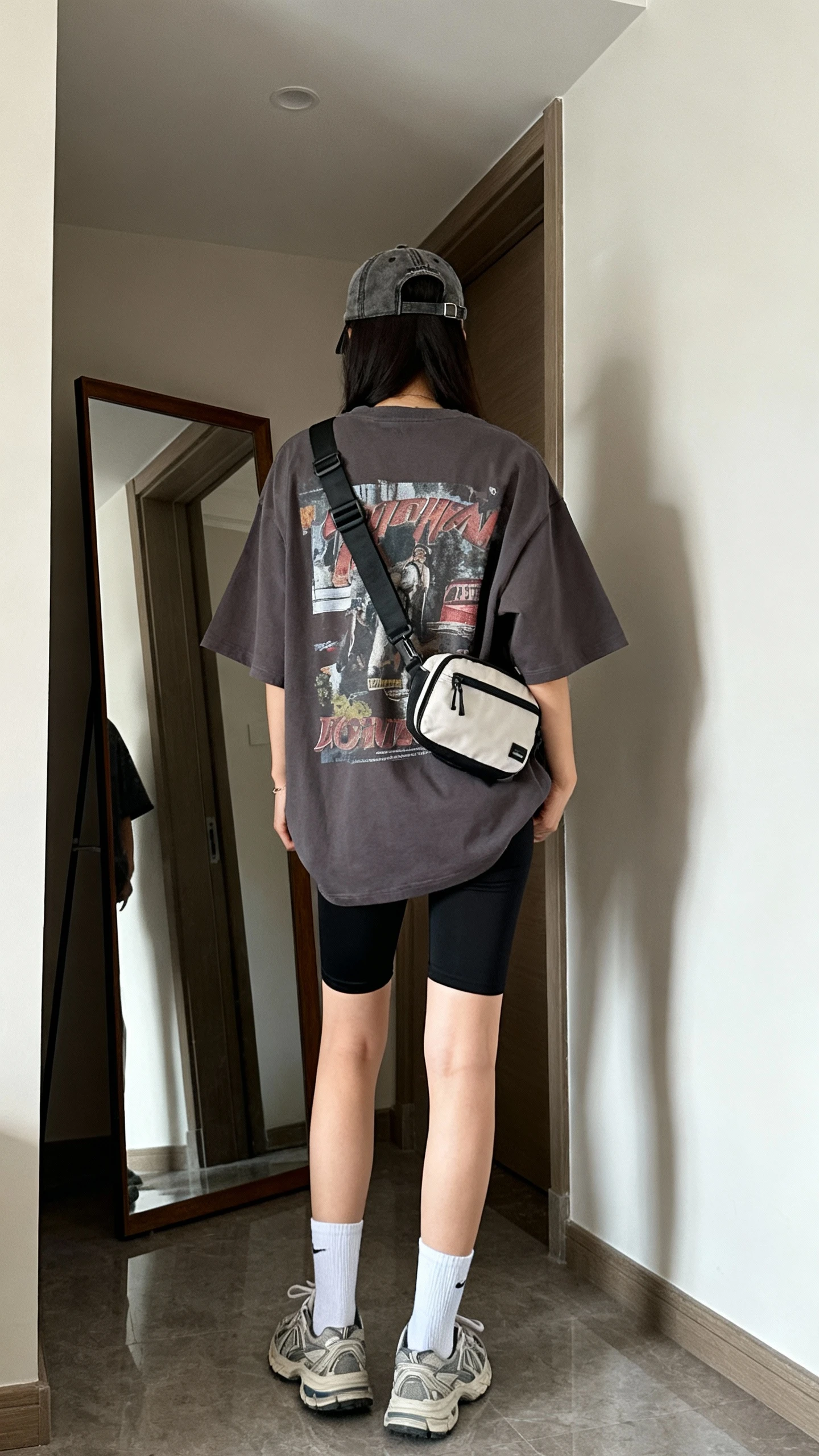 iPhone back view of a woman wearing an oversized graphic tee with black biker shorts, white crew socks, dad sneakers, a baseball cap, and a belt bag worn crossbody, face not visible, hallway full-length mirror, morning light, iPhone photo quality.