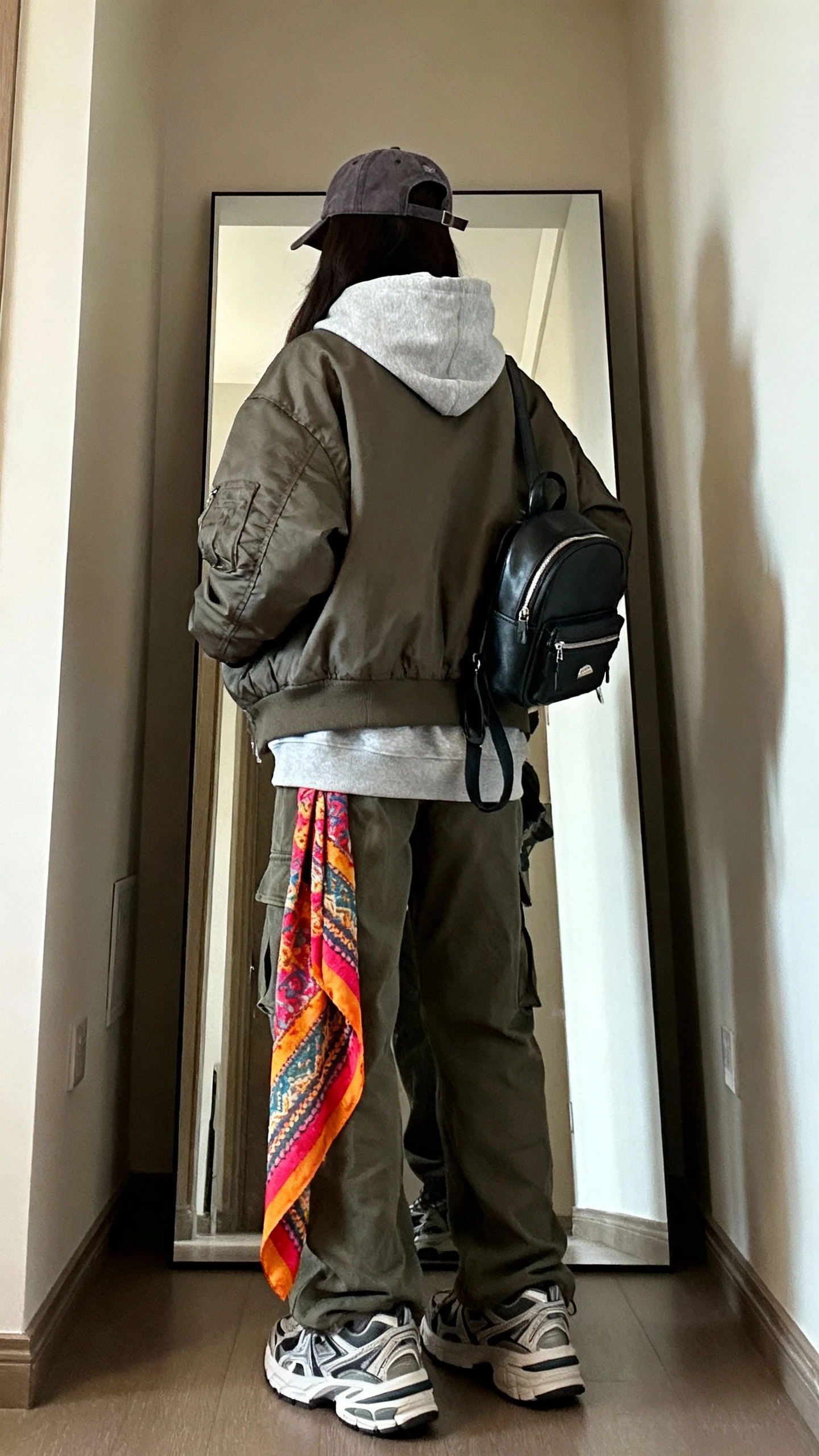 iPhone back view of a woman wearing an oversized hoodie layered under a boxy bomber jacket, cargo pants, chunky sneakers, a vibrant patterned scarf tied long, baseball cap, and mini backpack, face not visible, hallway mirror, indoor daylight, casual iPhone photo.