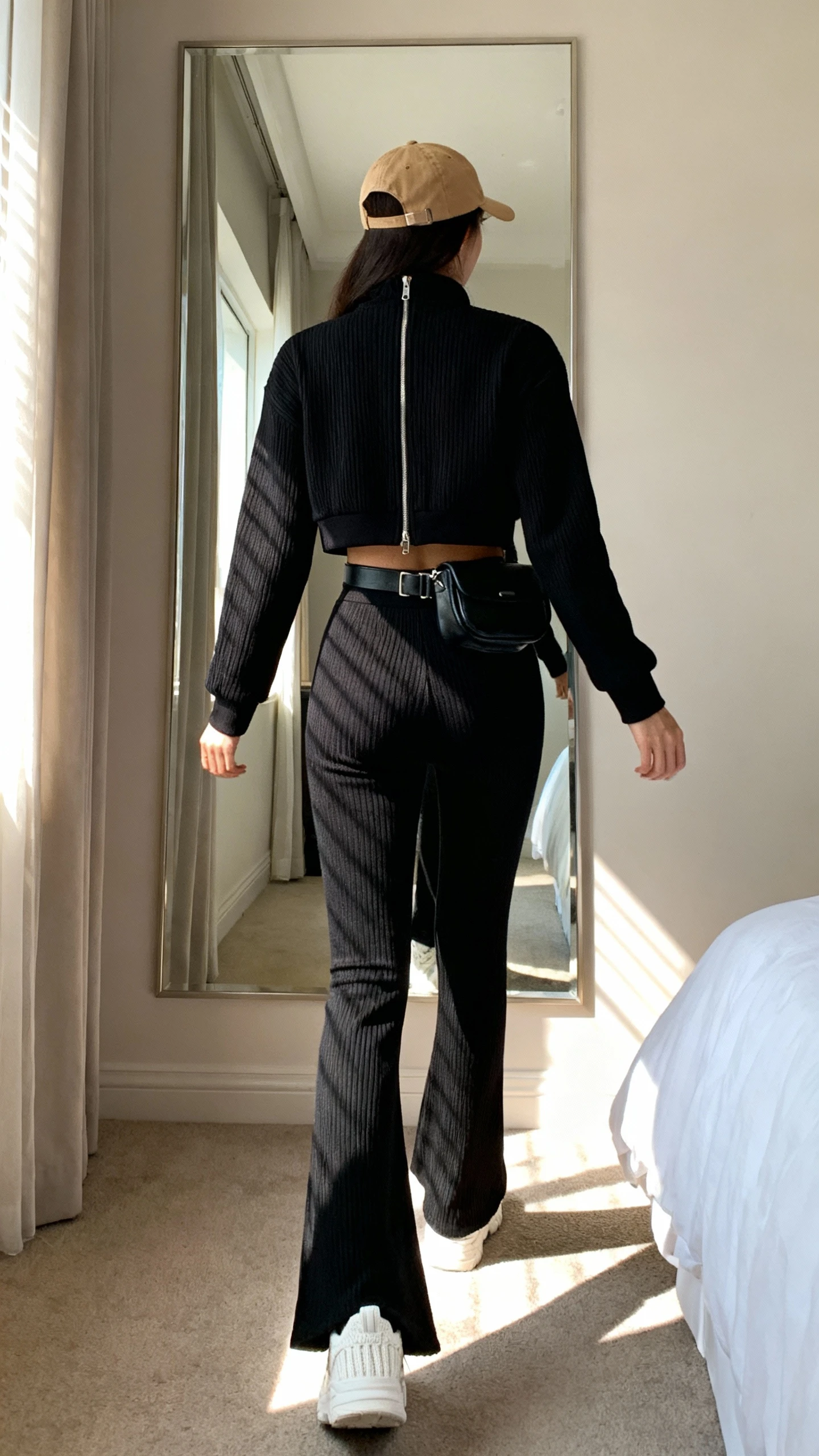iPhone back view of a woman wearing crisp white knit sneakers, a matching black ribbed knit set (cropped zip-up track jacket and high-waist flare leggings), a lightweight camel baseball cap, and a small black belt bag, face not visible, walking pose in bedroom mirror with afternoon window light, iPhone photo quality.