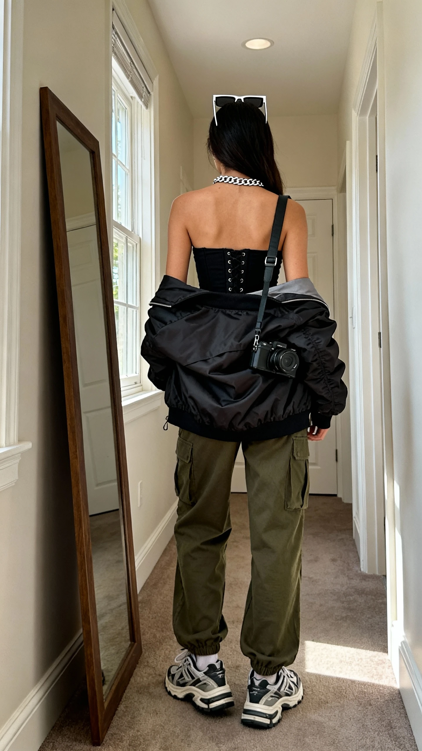 iPhone back view of a woman wearing elevated streetwear: black corset top, olive cargo pants, oversized bomber jacket draped over shoulders, chunky sneakers, silver chain necklace, rectangular sunglasses on head, and a crossbody camera bag, face not visible, hallway full-length mirror, afternoon window light, iPhone photo quality.