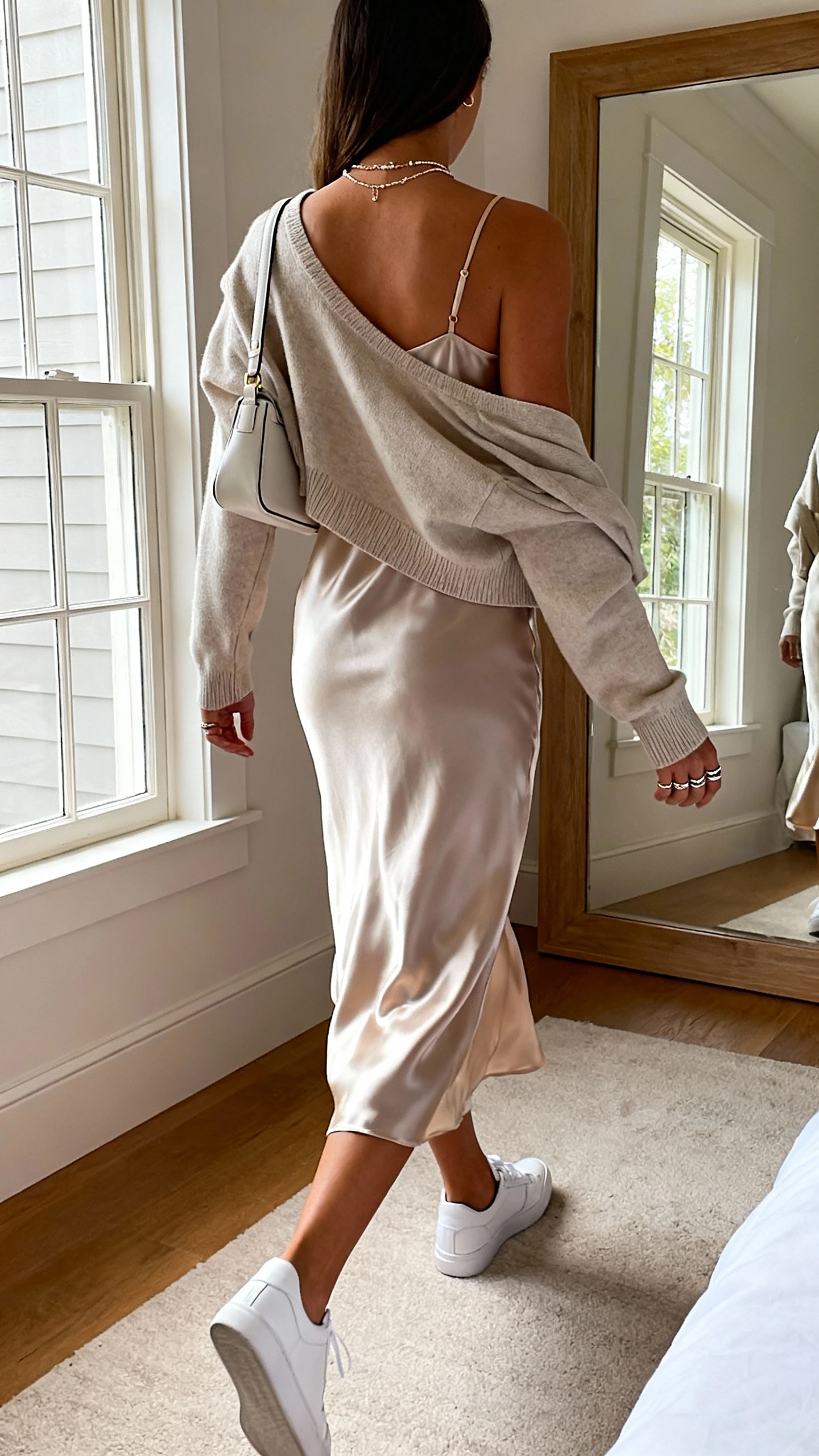 iPhone back-view photo of a woman in a satin/silky midi slip dress, white minimalist sneakers with a sleek shape, a cropped cardigan or boyfriend blazer or leather jacket draped over shoulders, a small shoulder bag or chain-strap mini bag, layered delicate necklaces and stacked rings, face not visible, walking pose toward a bedroom mirror, soft afternoon window light, iPhone photo quality.