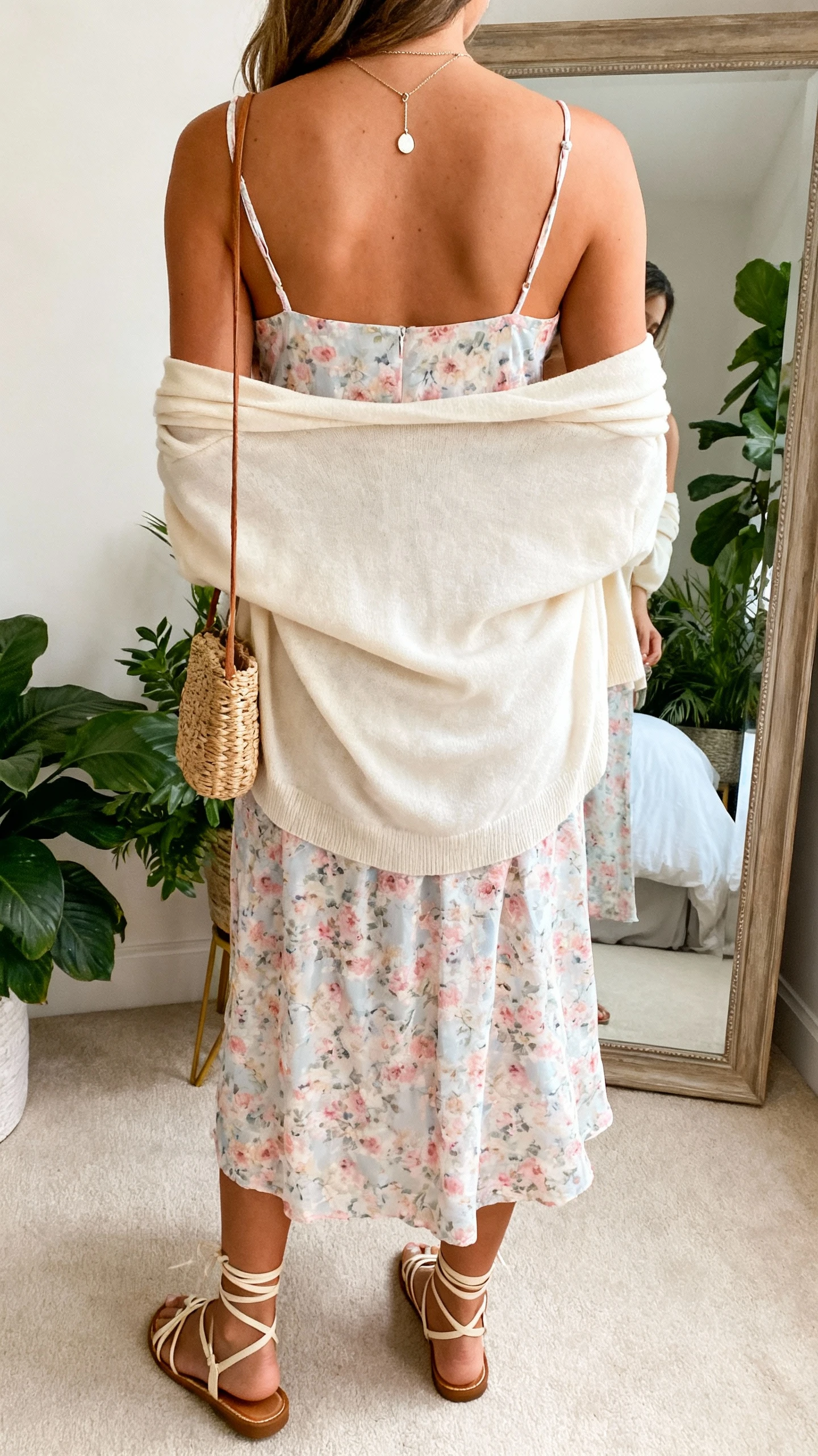 iPhone back-view photo of a woman wearing a pastel floral slip dress with a soft cream cardigan draped over shoulders, strappy sandals, delicate pendant necklace, and a small woven bag; face not visible, back view, bedroom mirror with plants, gentle morning light, iPhone photo quality.