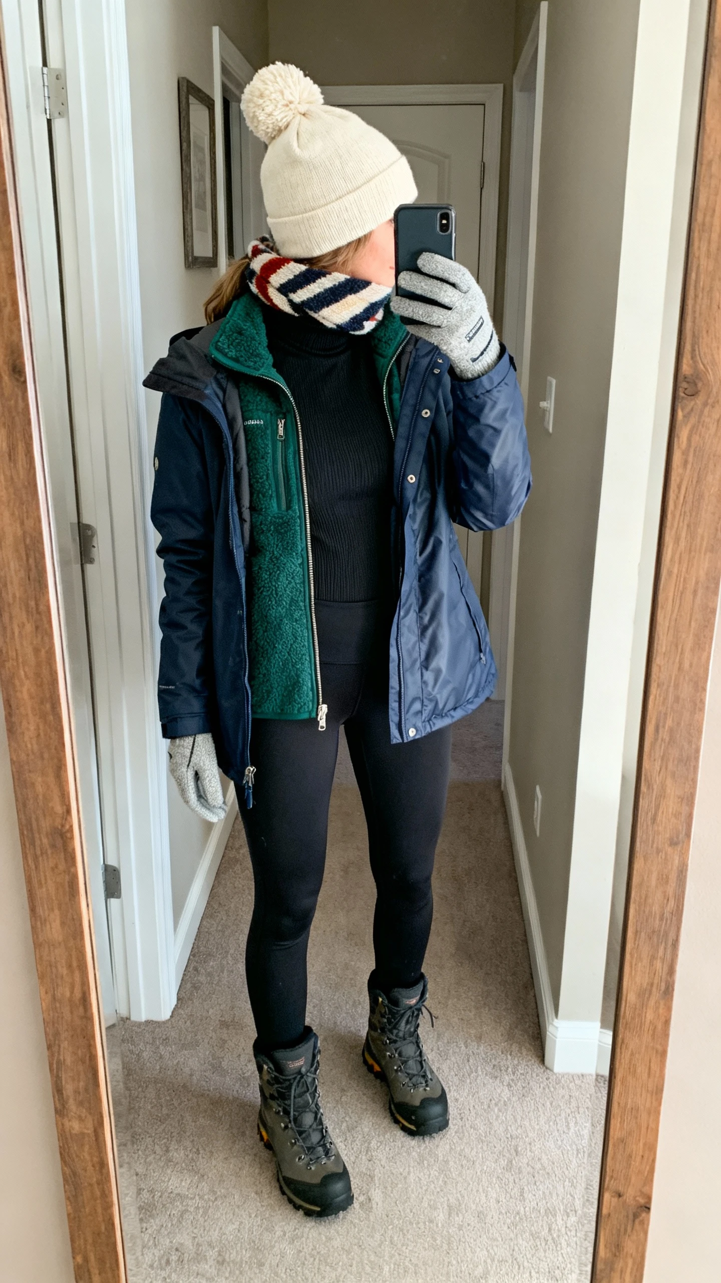 iPhone back-view selfie in a hallway mirror of a woman wearing a black thermal turtleneck, a forest-green sherpa zip-up, a navy water-resistant parka, insulated black leggings, weatherproof hiker boots, a chunky cream beanie with touchscreen gloves, and a playful striped scarf, face not visible, indoor afternoon light, iPhone photo quality.