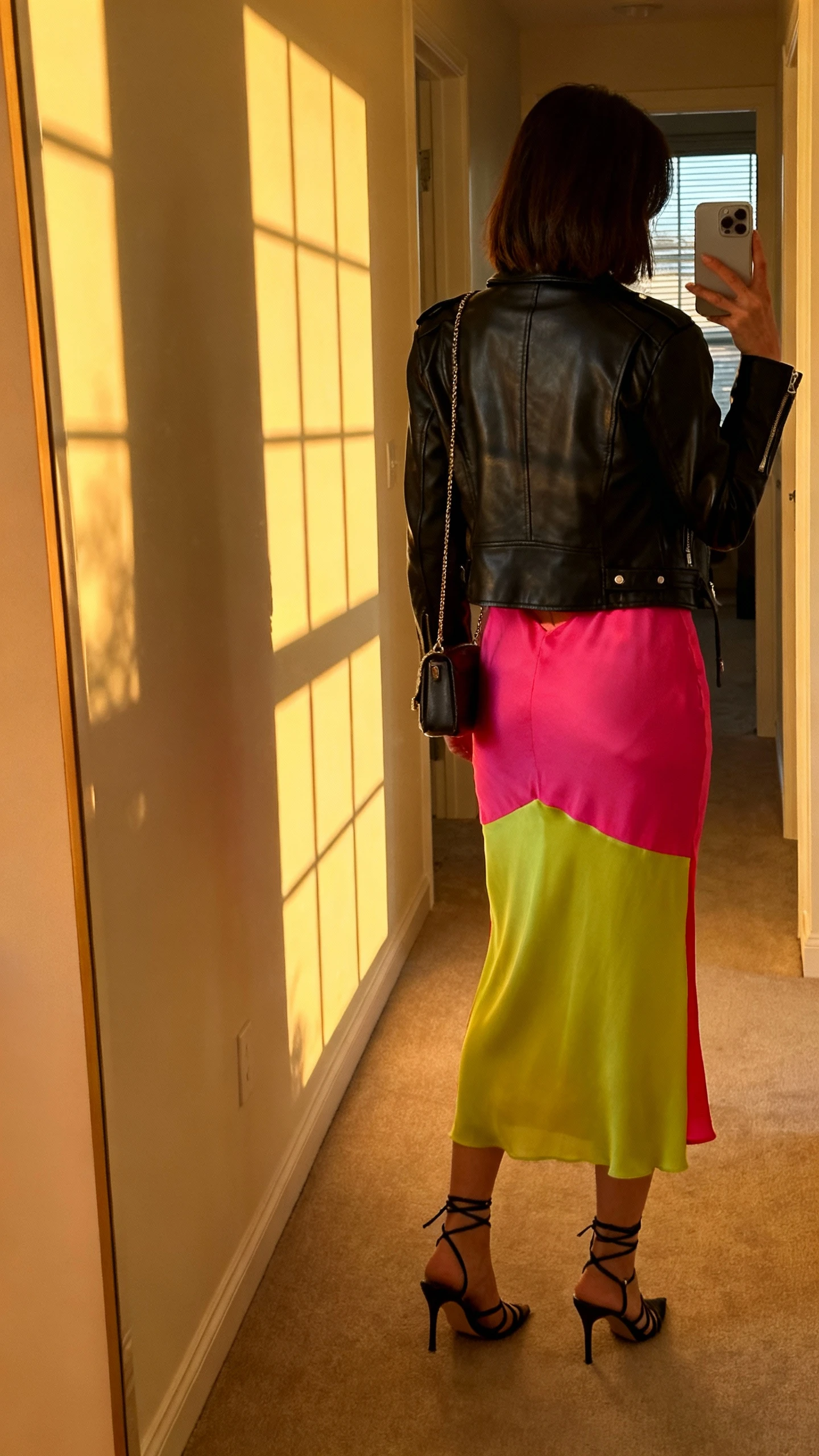 iPhone back-view selfie of a woman wearing a bright color-pop slip dress layered with a black leather moto jacket, strappy heels, and a small shoulder bag, face not visible, golden-hour window light in a hallway mirror, iPhone photo quality.