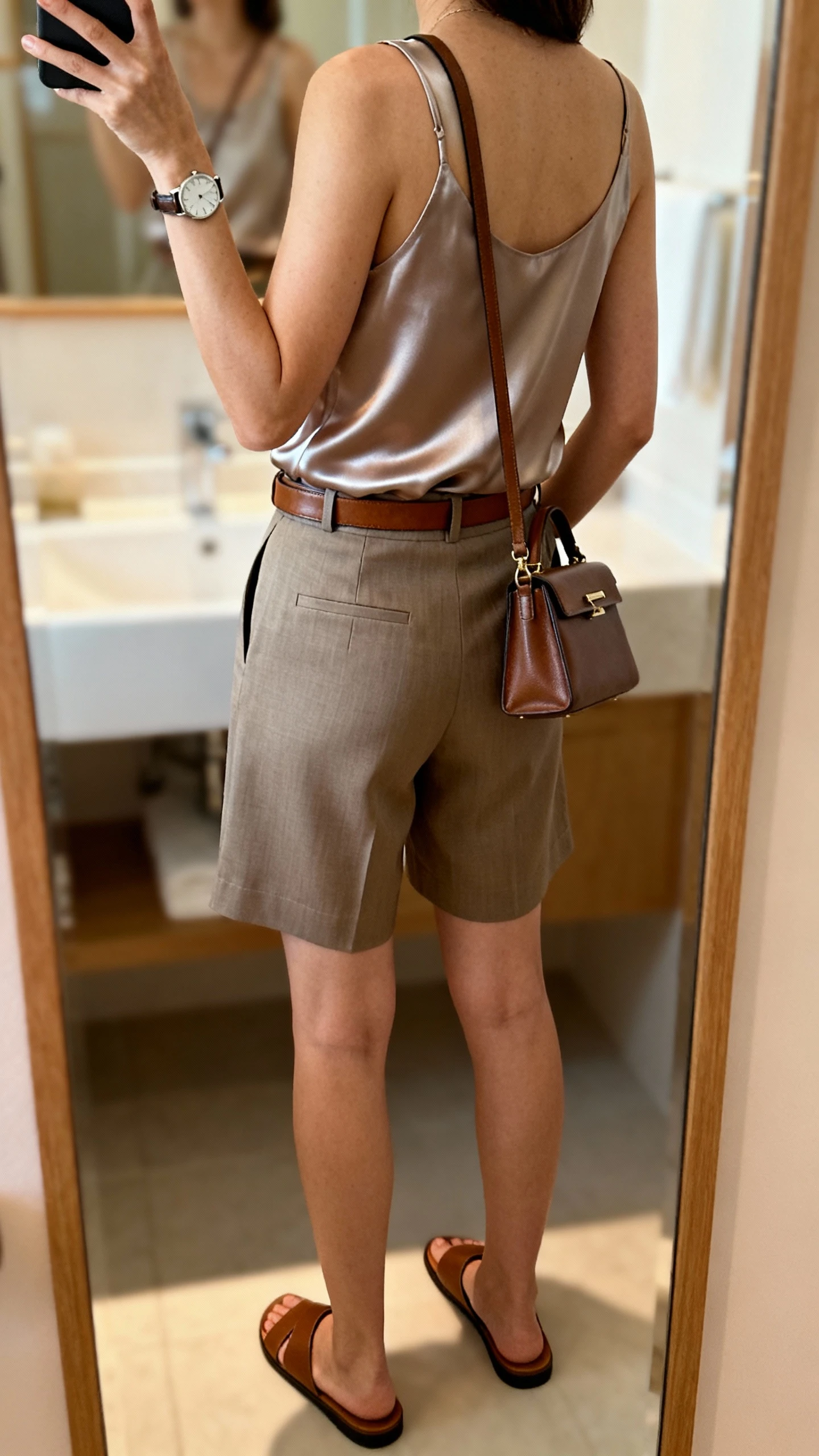 iPhone back-view selfie of a woman wearing tailored high-waist shorts, a silk tank top tucked in, leather belt, minimalist leather slides, structured crossbody bag, sleek watch, face not visible, bathroom mirror with soft indoor lighting, iPhone photo quality