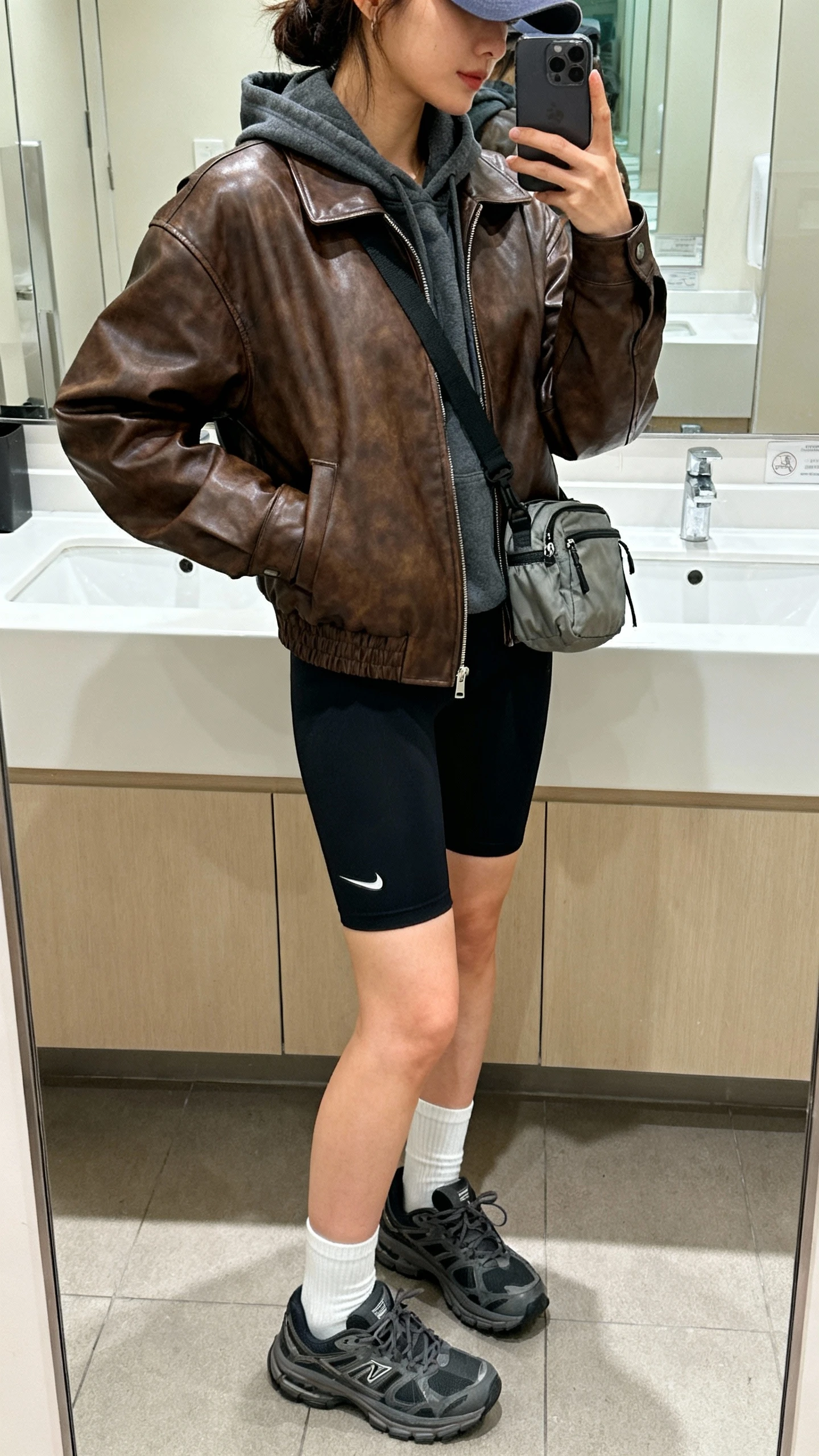 iPhone bathroom mirror selfie of a woman wearing a brown leather bomber jacket with a charcoal hoodie, black bike shorts, chunky dad sneakers, white crew socks, mini nylon shoulder bag, baseball cap, face not visible, side angle crop at shoulders, indoor lighting, iPhone photo quality
