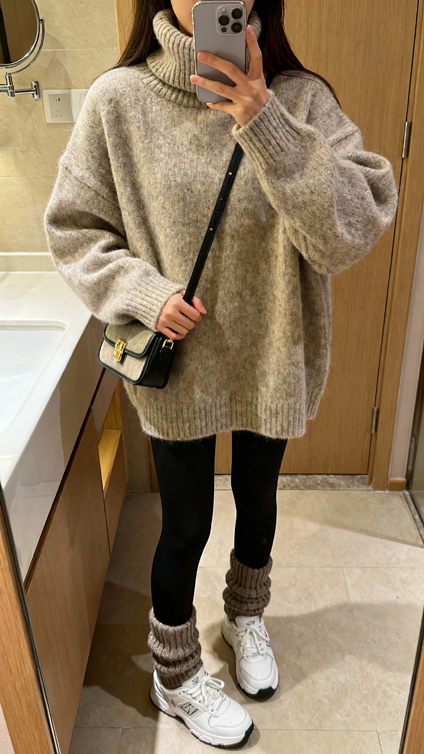 iPhone bathroom mirror selfie of a woman wearing an oversized turtleneck sweater with black leggings, chunky socks and white sneakers, small crossbody bag in hand, face not visible, cropped at shoulders, warm indoor lighting, iPhone photo quality.