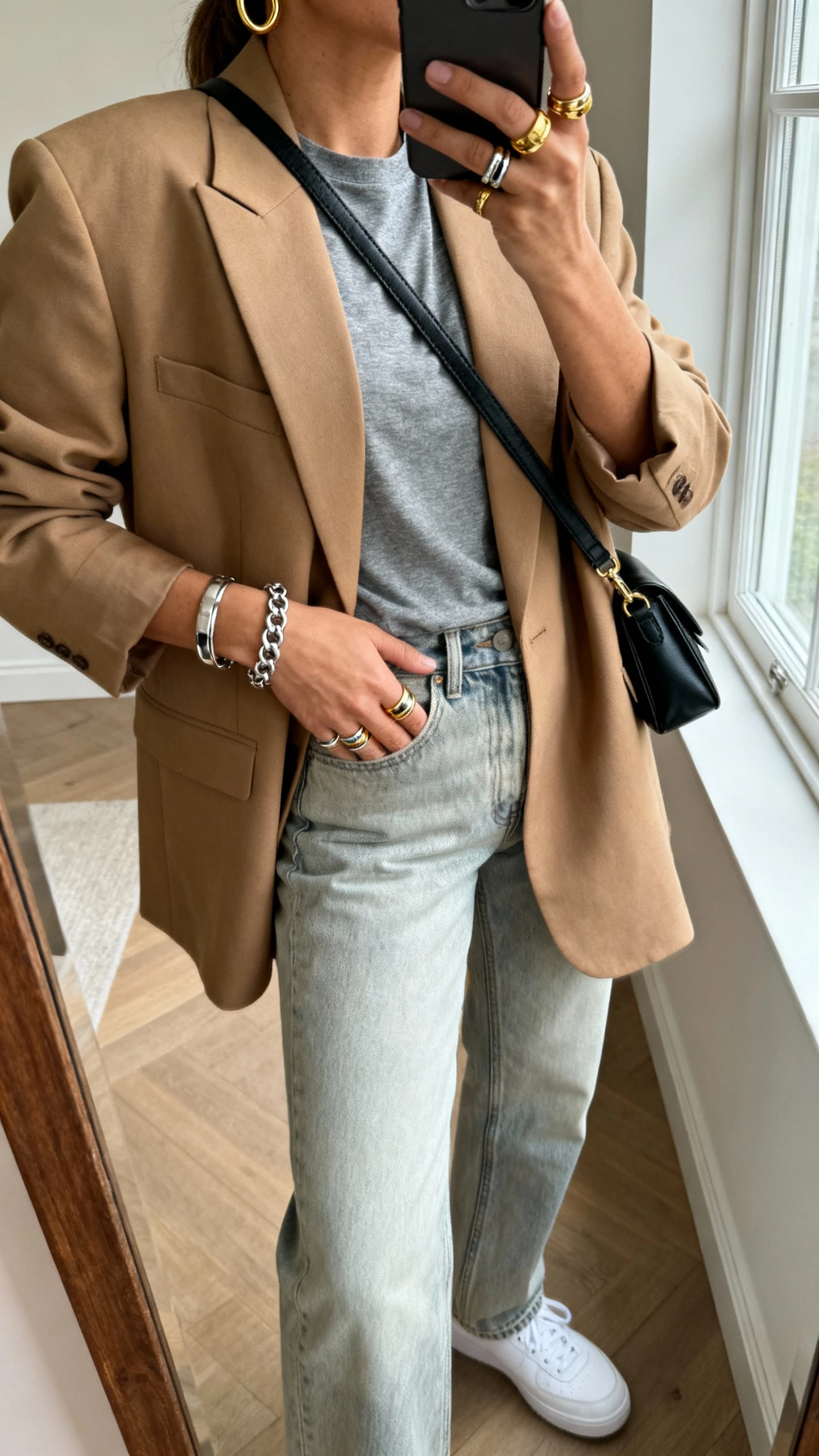iPhone casual mirror selfie of a woman wearing a grey tee tucked into light-wash straight jeans, oversized camel blazer, white sneakers, mixed metals jewelry (gold hoops, silver chain bracelet, gold-silver layered rings), and a black crossbody bag, face not visible, side angle, natural window light, iPhone photo quality