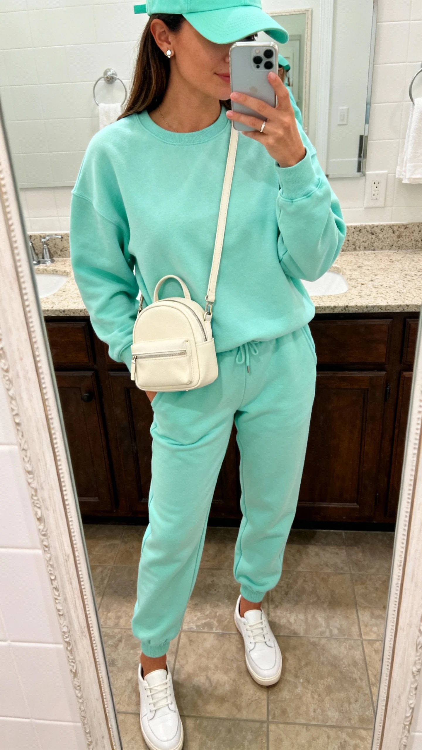 iPhone casual mirror selfie of a woman wearing a mint sweatshirt with matching mint joggers, white low-profile leather sneakers, a cream mini backpack, a mint baseball cap, and simple stud earrings, face not visible, cropped at neck, bathroom mirror, natural indoor lighting, iPhone photo quality.
