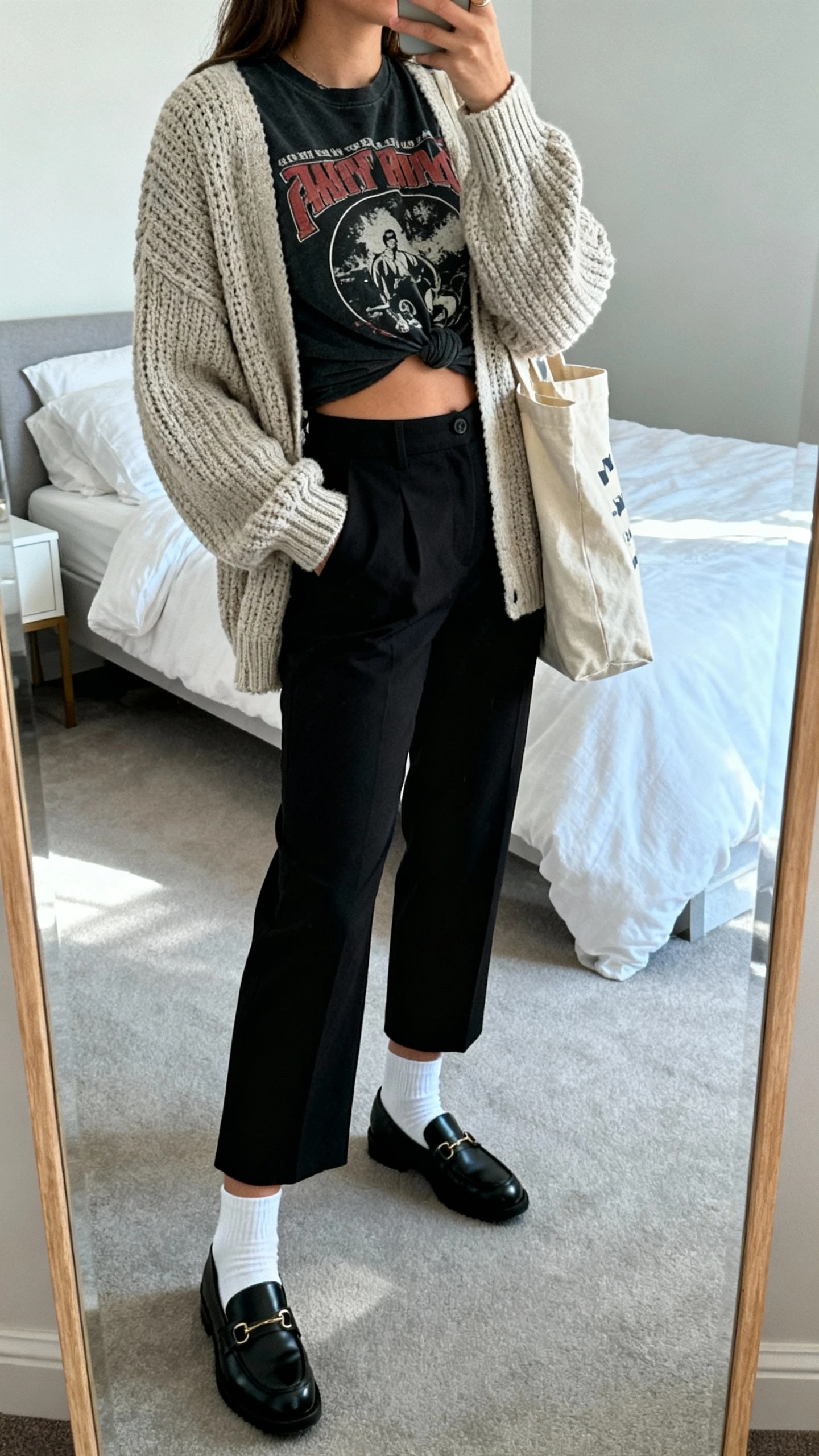 iPhone casual mirror selfie of a woman wearing black cropped trousers, a vintage band tee half-tucked, a slouchy fisherman cardigan, black loafers with white socks, and a canvas tote, face not visible, side angle in bedroom mirror with cool morning light, iPhone photo quality.