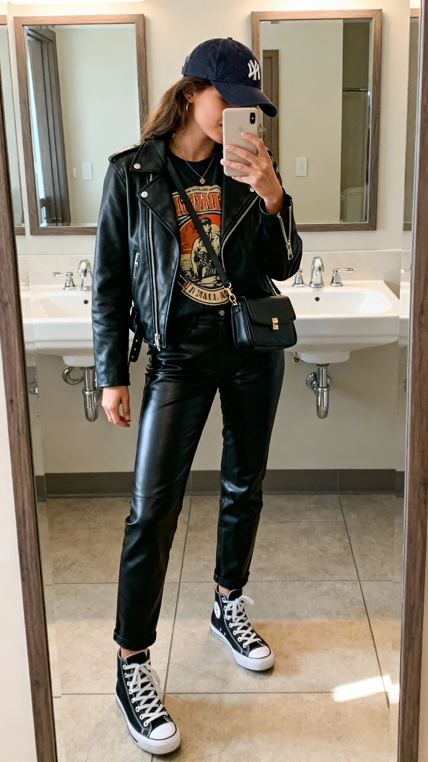 iPhone full-length mirror selfie of a woman wearing black leather pants, a vintage-style graphic tee, a black leather moto jacket, high-top sneakers, a baseball cap, and a small crossbody bag; face not visible, side angle, bathroom mirror, indoor morning light, iPhone photo quality.