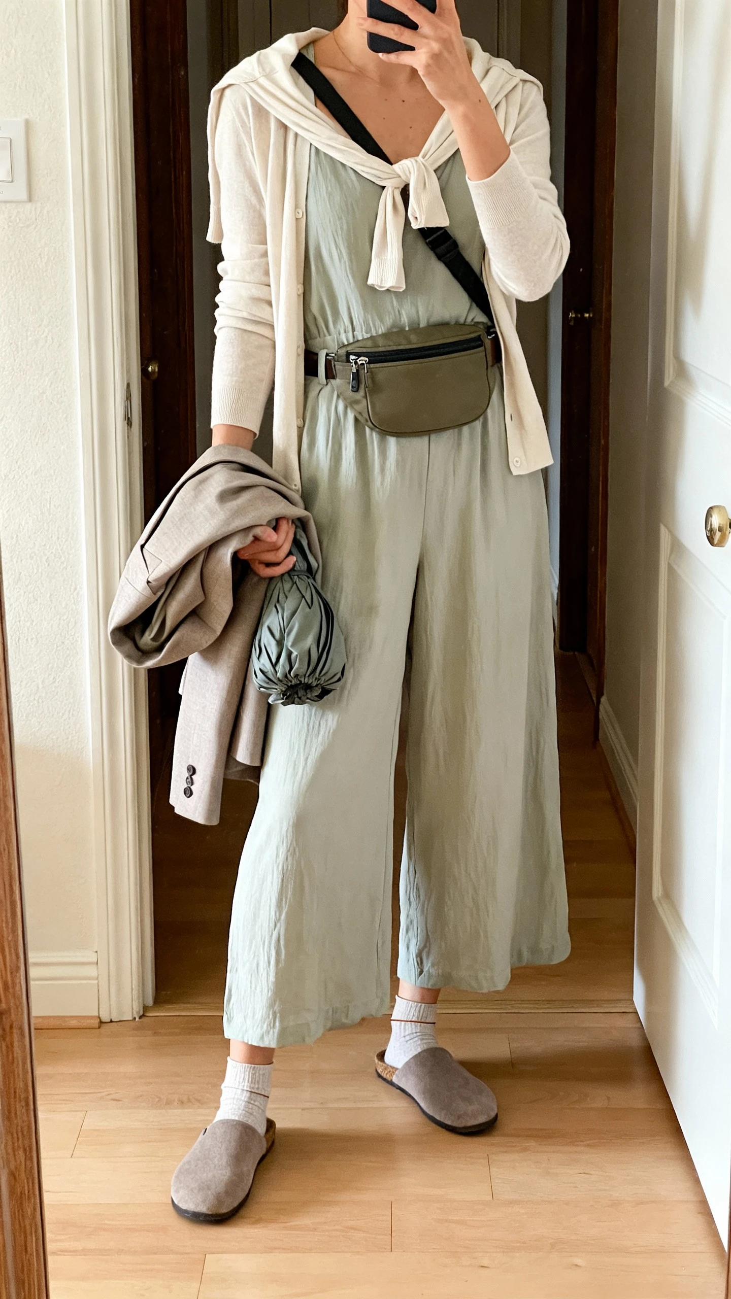 iPhone mirror selfie of a woman in a breezy wide-leg jumpsuit with a light cardigan tied at the shoulders and a packable blazer in hand, slip-on mules, belt bag worn crossbody, and thin socks for security, face not visible, cropped at neck, entryway mirror with natural morning light, iPhone photo quality.