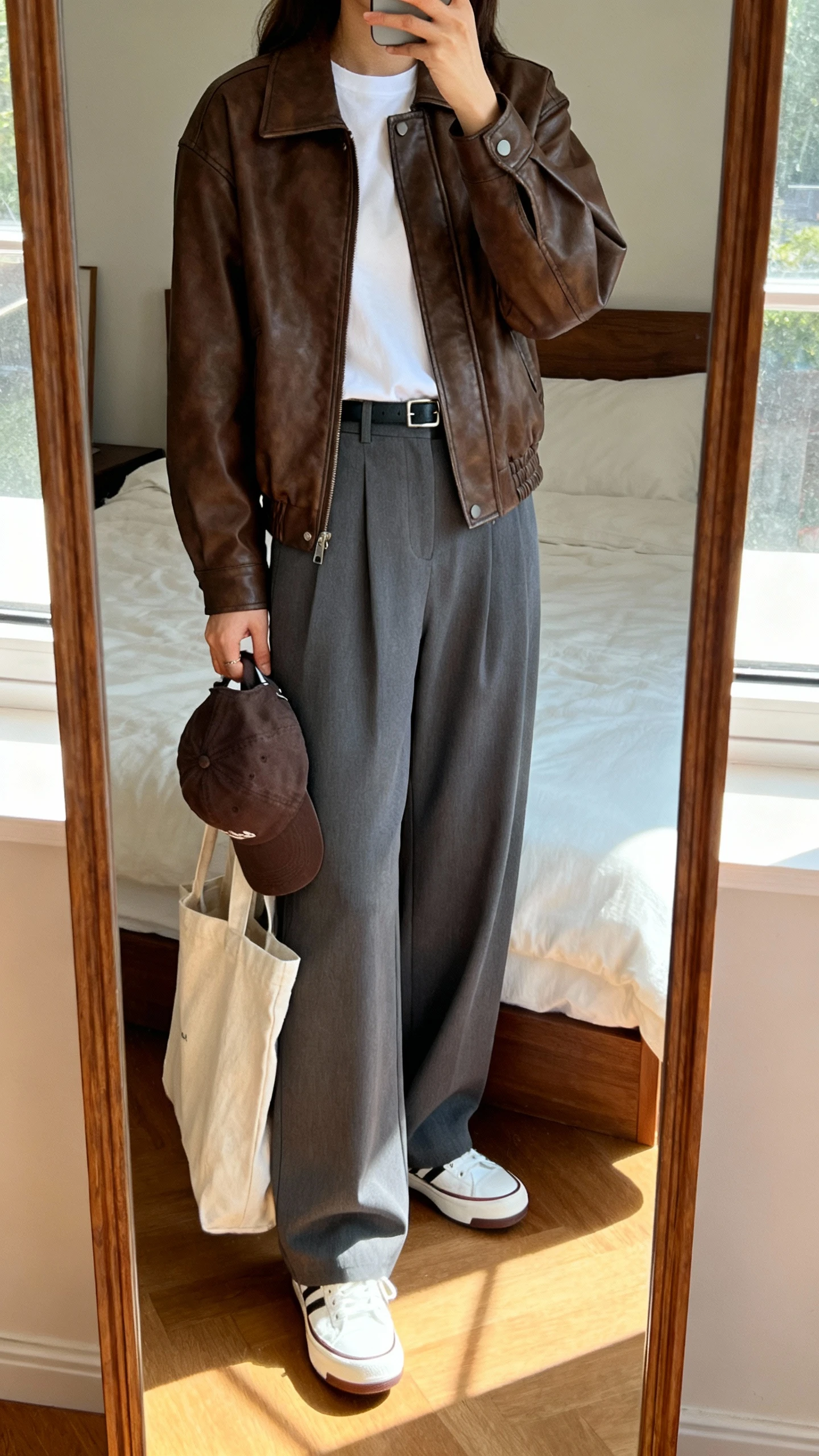 iPhone mirror selfie of a woman in a brown leather jacket, gray wide-leg trousers, white tee tucked in, white retro sneakers, baseball cap tucked in hand, and a canvas tote, face not visible, cropped at shoulders, bedroom mirror, bright window light, iPhone photo quality.
