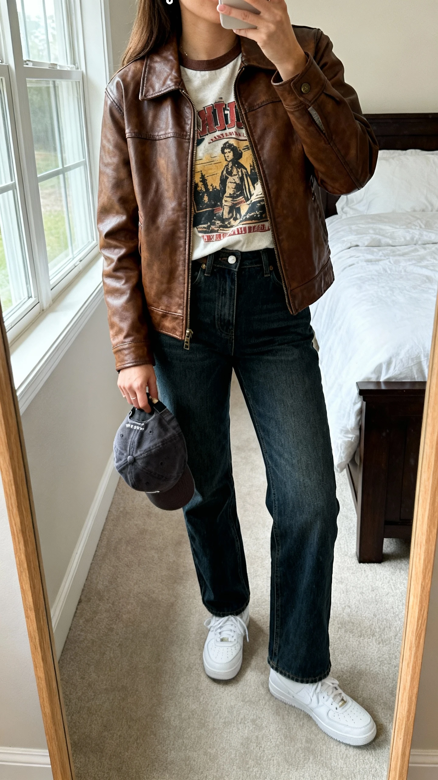 iPhone mirror selfie of a woman in a brown leather jacket with a vintage graphic tee tucked into straight-leg jeans, white sneakers, baseball cap in hand, simple studs, face not visible, cropped at shoulders, bedroom mirror, natural window light, iPhone photo quality
