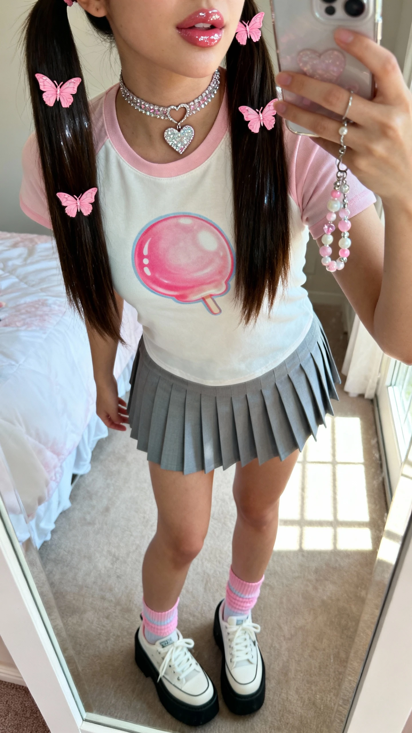 iPhone mirror selfie of a woman in a Bubblegum Pop Princess look: glossy high pigtails with pink butterfly clips, baby tee with a bubblegum graphic, pleated mini skirt, platform sneakers with pastel ankle socks, sparkly heart choker, beaded phone charm, glossy lip look implied, face not visible, cropped at neck, bedroom mirror with natural window light, iPhone photo quality