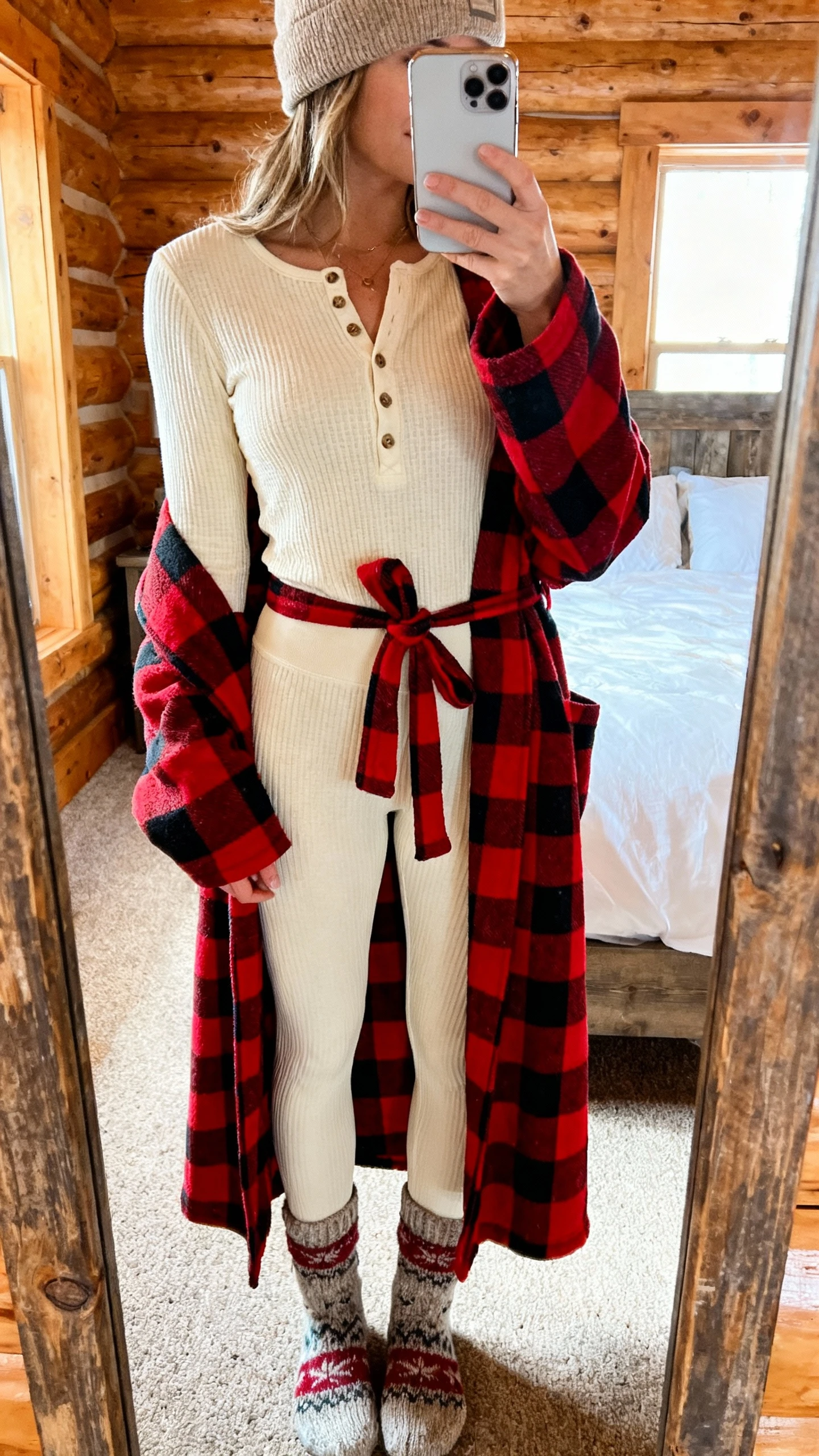 iPhone mirror selfie of a woman in a cabin-cozy thermal set: waffle-knit cream henley top and matching thermal leggings, red Buffalo-check flannel robe tied at waist, wool cabin socks, beanie; face not visible, cropped at shoulders, rustic bedroom mirror with soft morning light, iPhone photo quality.