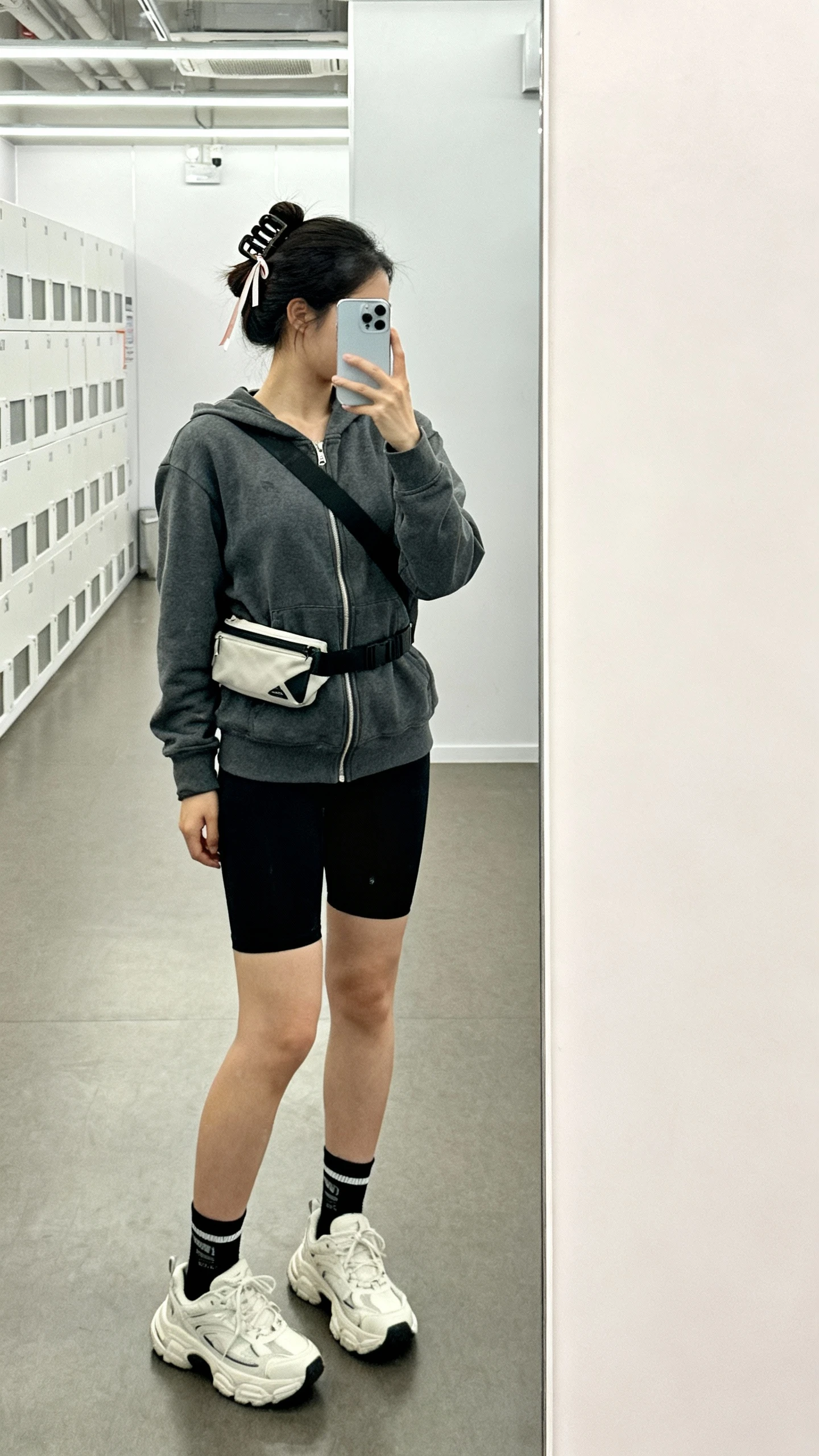 iPhone mirror selfie of a woman in a charcoal zip-up hoodie, black bike shorts, chunky white sneakers, crossbody belt bag, crew socks, and hair in a mega claw clip with a small ribbon detail threaded through; face not visible, gym locker-room mirror, bright indoor lighting, iPhone photo quality.