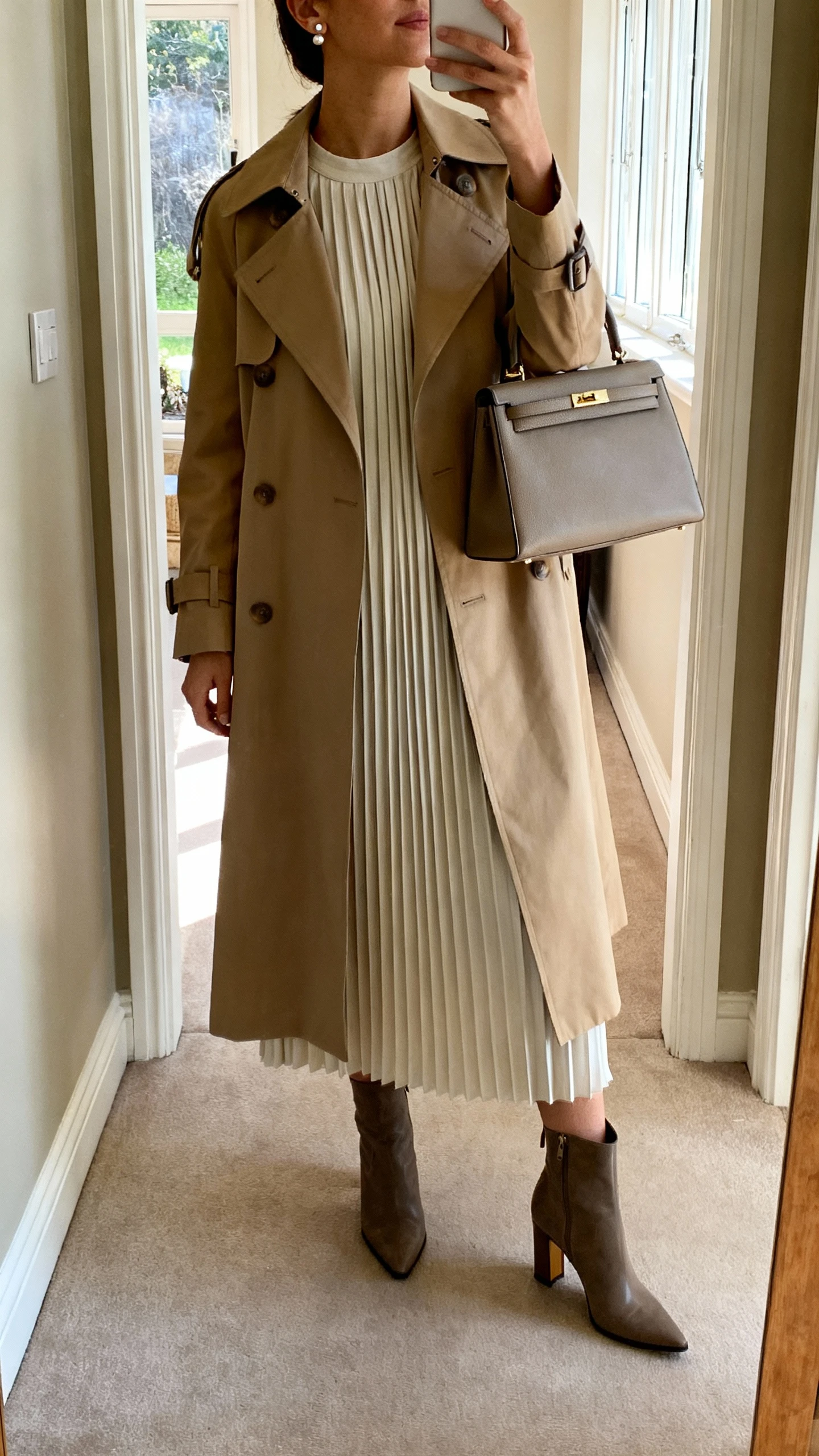 iPhone mirror selfie of a woman in a column dress layered under a classic trench coat, heeled ankle boots, structured top-handle bag, subtle stud earrings, face not visible, cropped at neck, entryway mirror with soft afternoon window light, iPhone photo quality.