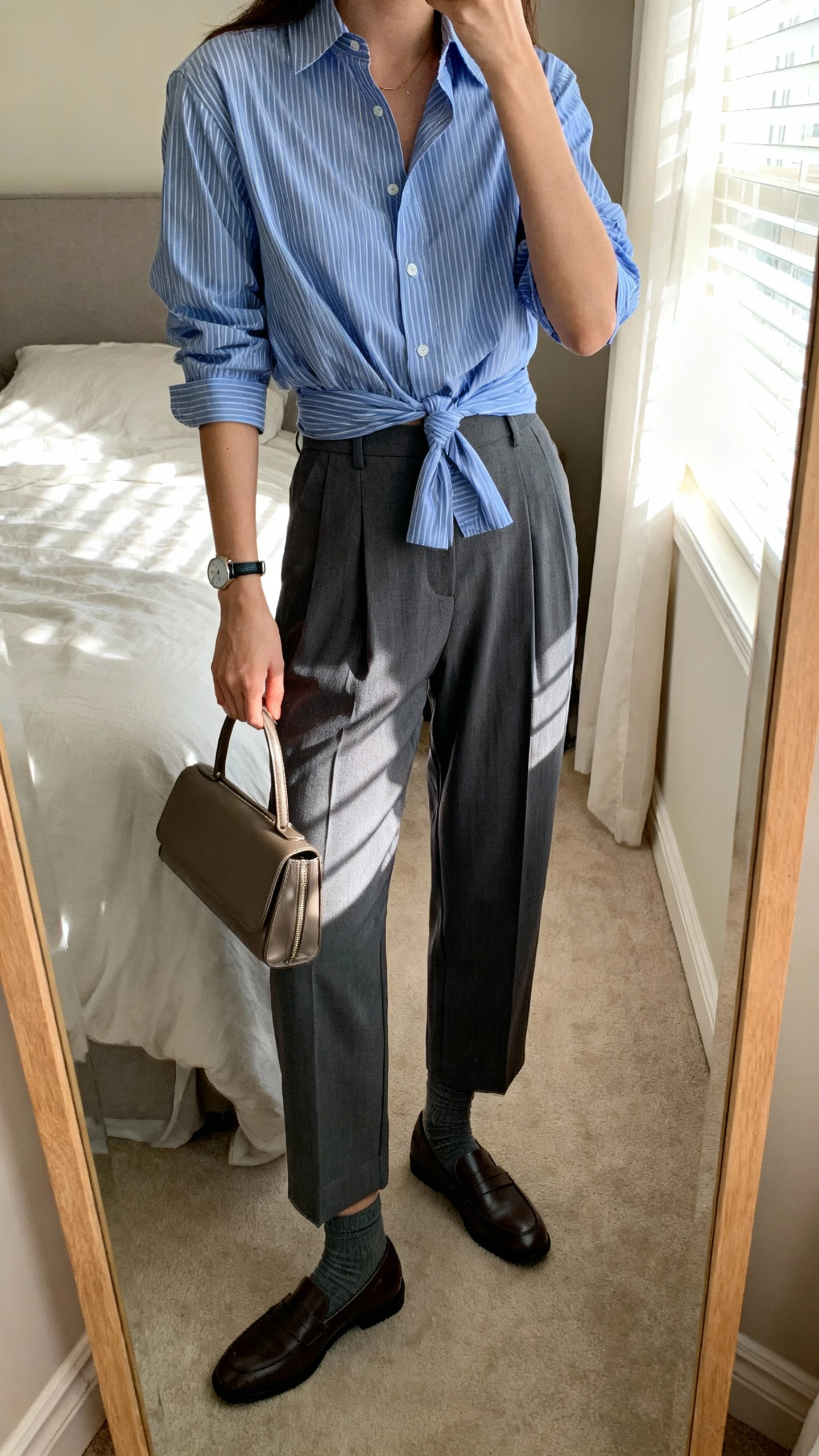 iPhone mirror selfie of a woman in a crisp blue striped button-down loosely tucked into relaxed pleated trousers in charcoal, loafers with slim socks, simple watch, and a slim leather tote, face not visible, cropped at shoulders, bedroom mirror with soft window light, iPhone photo quality.