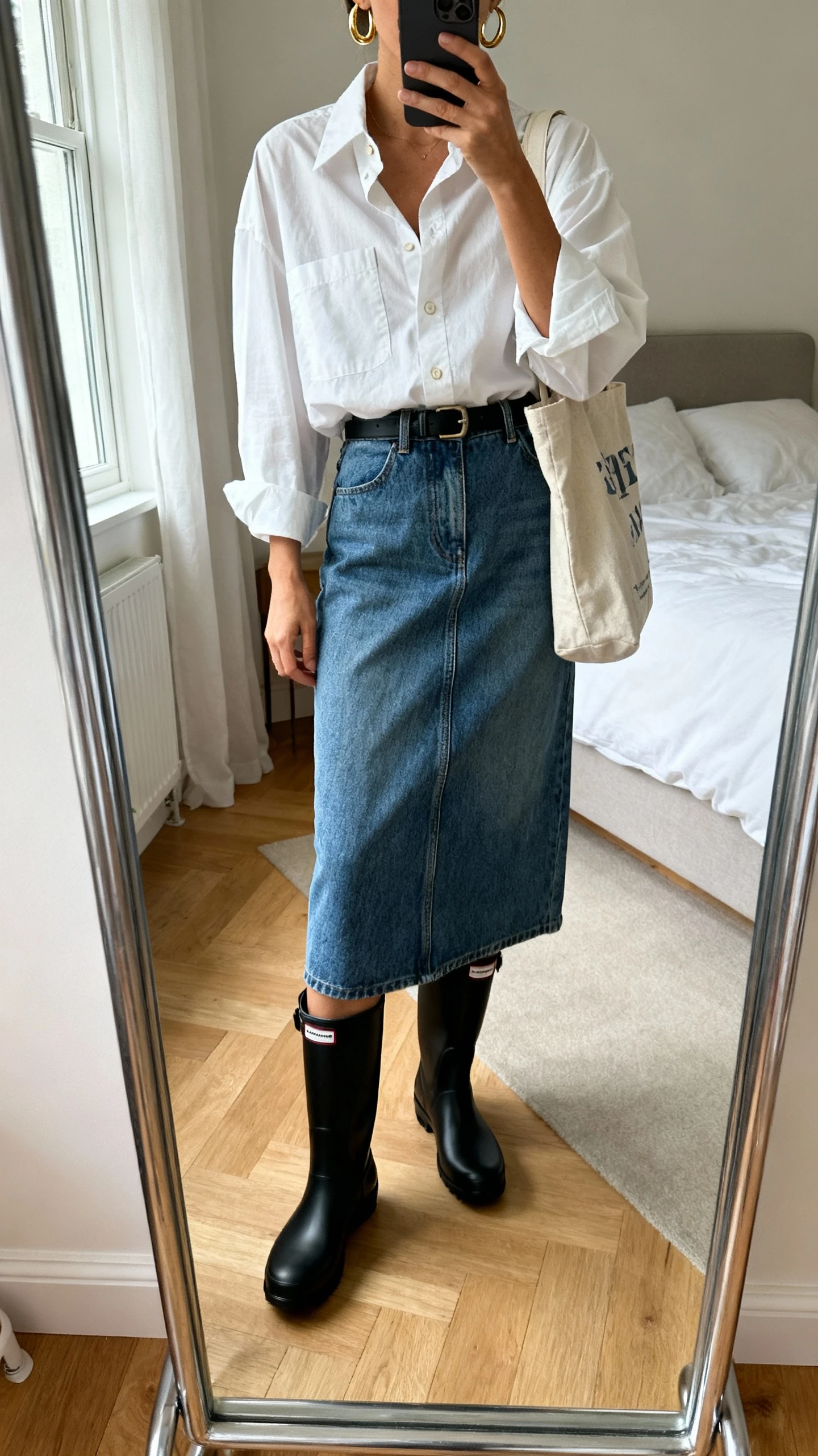 iPhone mirror selfie of a woman in a crisp oversized button-down tucked into a denim midi skirt, sleek black rain boots, minimal gold hoops, and a canvas tote, face not visible, cropped at shoulders, bedroom mirror with natural daylight, iPhone photo quality.