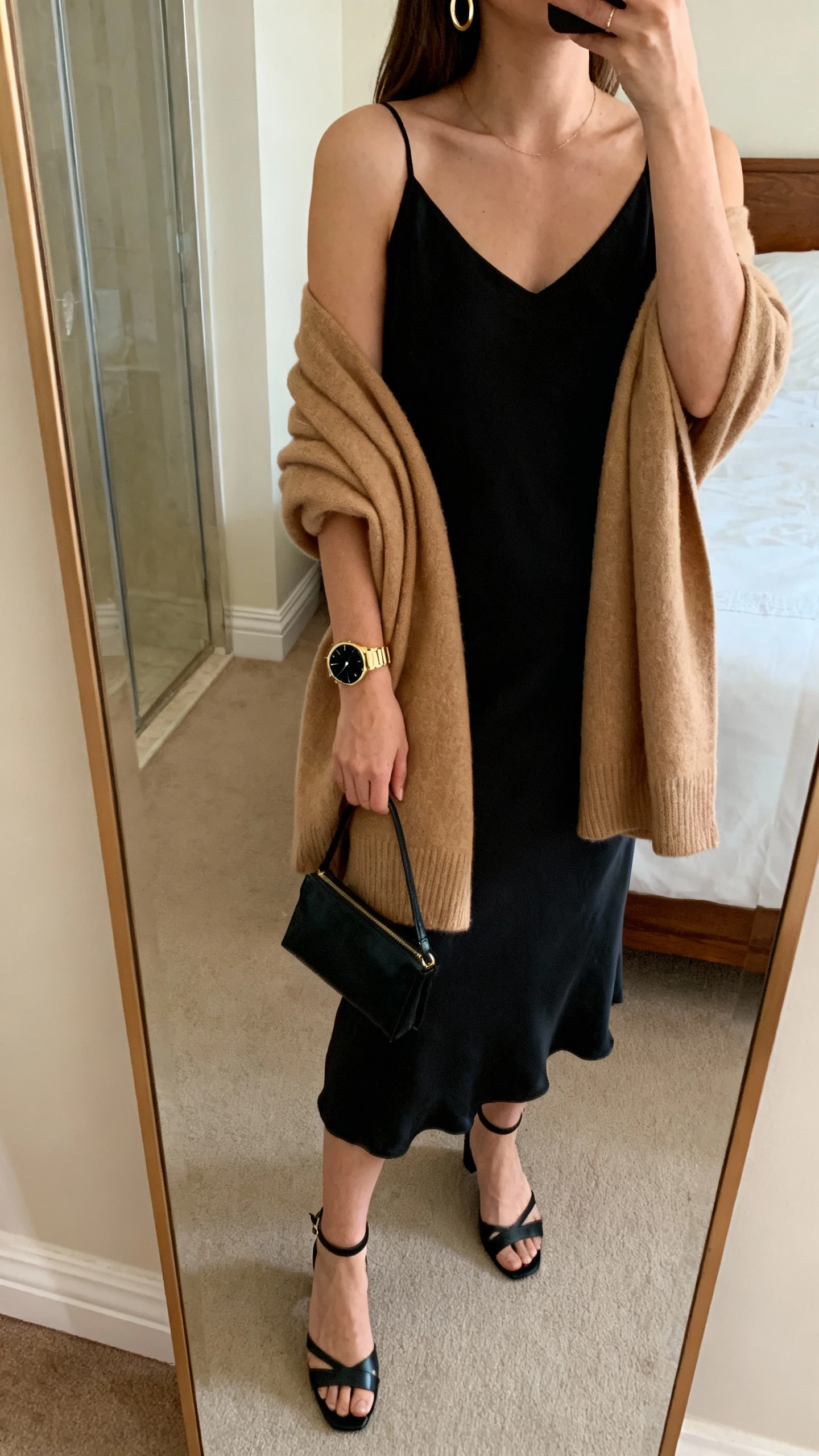 iPhone mirror selfie of a woman in a luxe-quiet evening look: black slip dress, camel cashmere wrap, strappy black heels, minimalist gold watch with black dial, delicate gold hoop earrings, small black clutch, face not visible, cropped at shoulders, bedroom mirror, warm indoor lighting, iPhone photo quality.