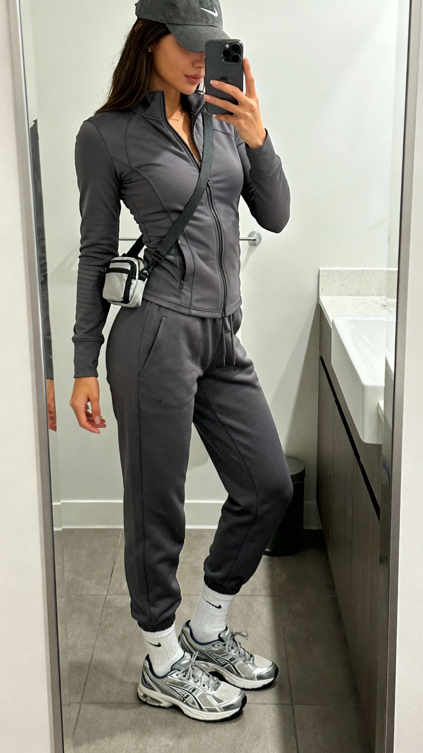 iPhone mirror selfie of a woman in a matching charcoal jogger set with a fitted zip-up track jacket, white crew socks, sleek running sneakers, baseball cap, and a small crossbody belt bag, face not visible, side angle, bathroom mirror, bright indoor lighting, iPhone photo quality.
