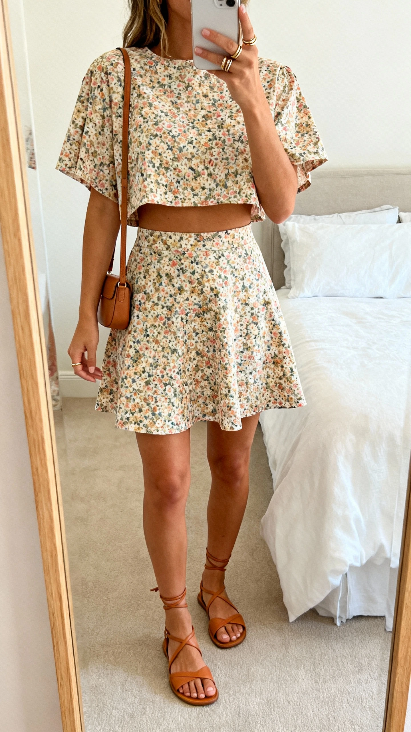 iPhone mirror selfie of a woman in a matching floral co-ord set (boxy short-sleeve crop top and A-line mini skirt), strappy tan sandals, small shoulder bag, and stacking rings; face not visible, cropped at neck, bedroom mirror, bright natural light, iPhone photo quality.