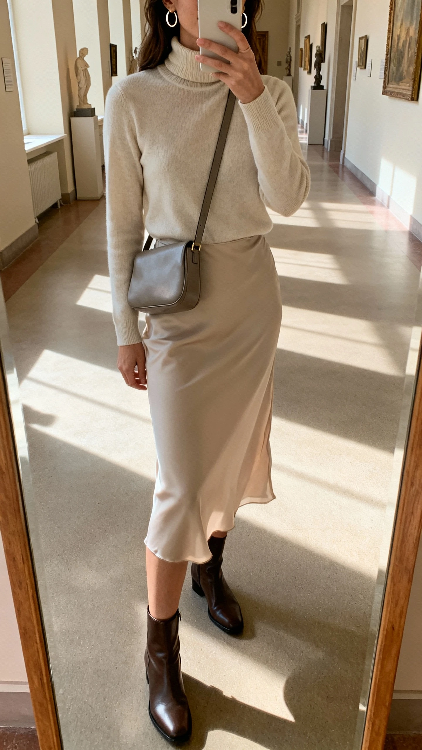 iPhone mirror selfie of a woman in a minimalist turtleneck, midi slip skirt, sleek ankle boots, thin hoop earrings, and a sleek crossbody bag worn across the chest, face not visible, cropped at shoulders, museum-like hallway mirror with soft morning light, iPhone photo quality.