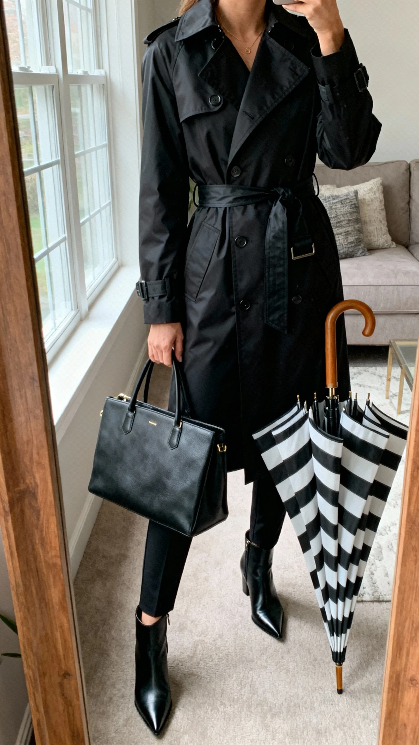 iPhone mirror selfie of a woman in a monochrome black outfit: sleek belted rain trench, slim trousers, pointed ankle boots, structured tote, bold striped umbrella as the statement, face not visible, cropped at shoulders, living room mirror, natural window light, iPhone photo quality.