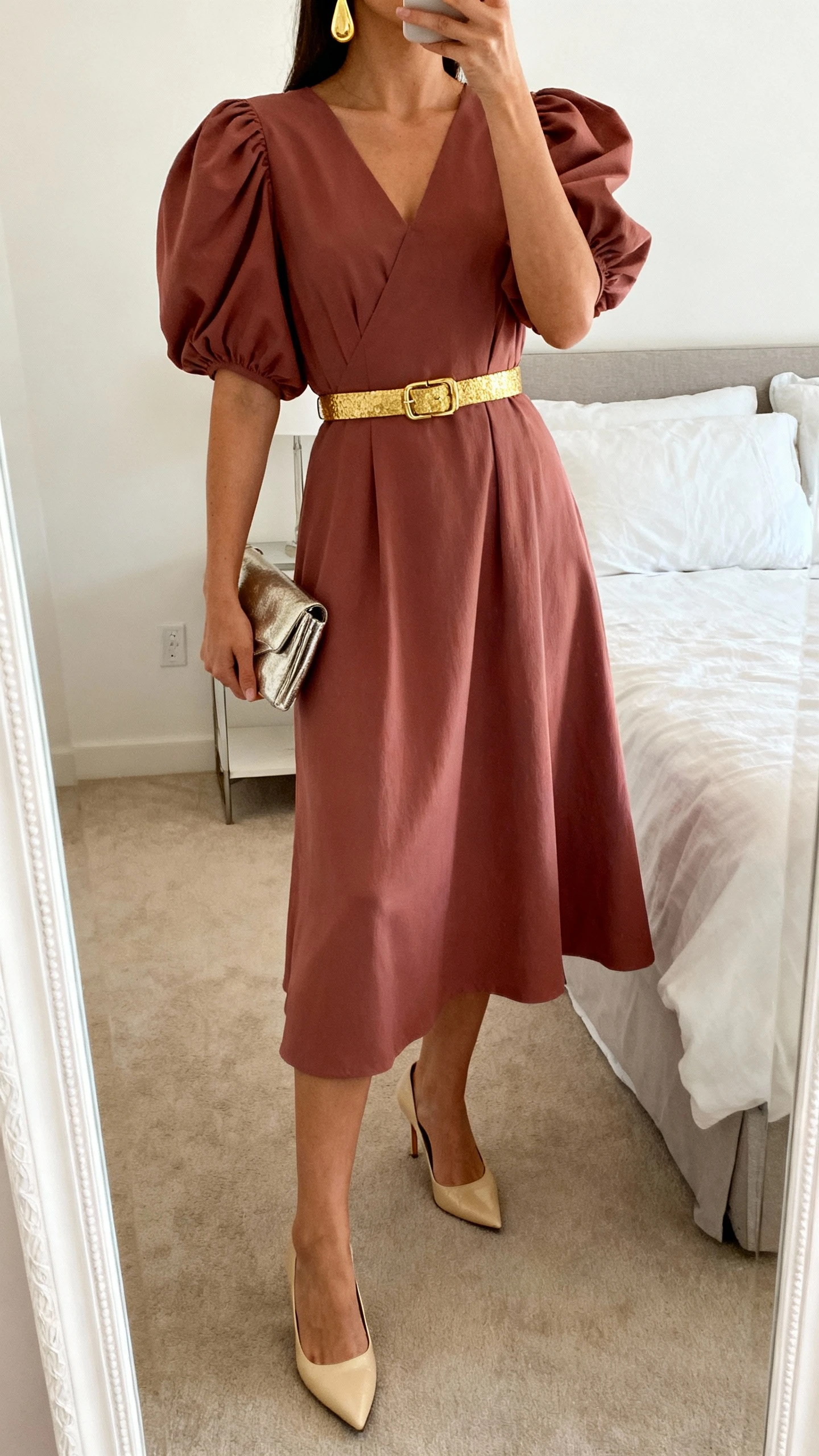 iPhone mirror selfie of a woman in a praline puff-sleeve midi dress with gilded accents (gold waist belt and gold drop earrings), champagne heels and a small metallic clutch, face not visible, cropped at neck, full-length mirror in bedroom, soft indoor lighting, iPhone photo quality.