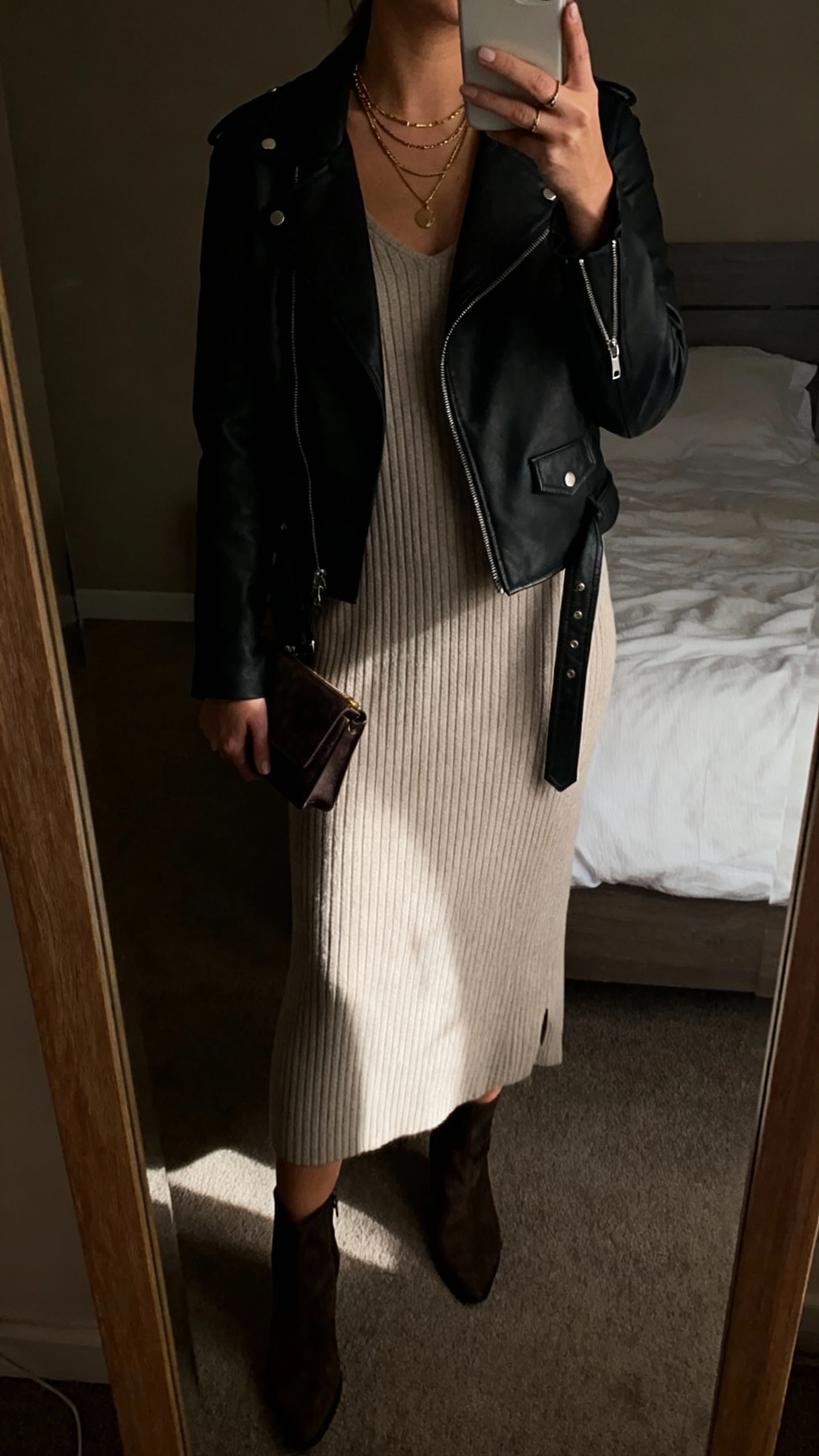 iPhone mirror selfie of a woman in a ribbed knit midi dress layered with a black moto jacket, ankle boots, a small clutch, and layered necklaces, face not visible, cropped at neck, dim bedroom mirror, warm indoor light, iPhone photo quality.