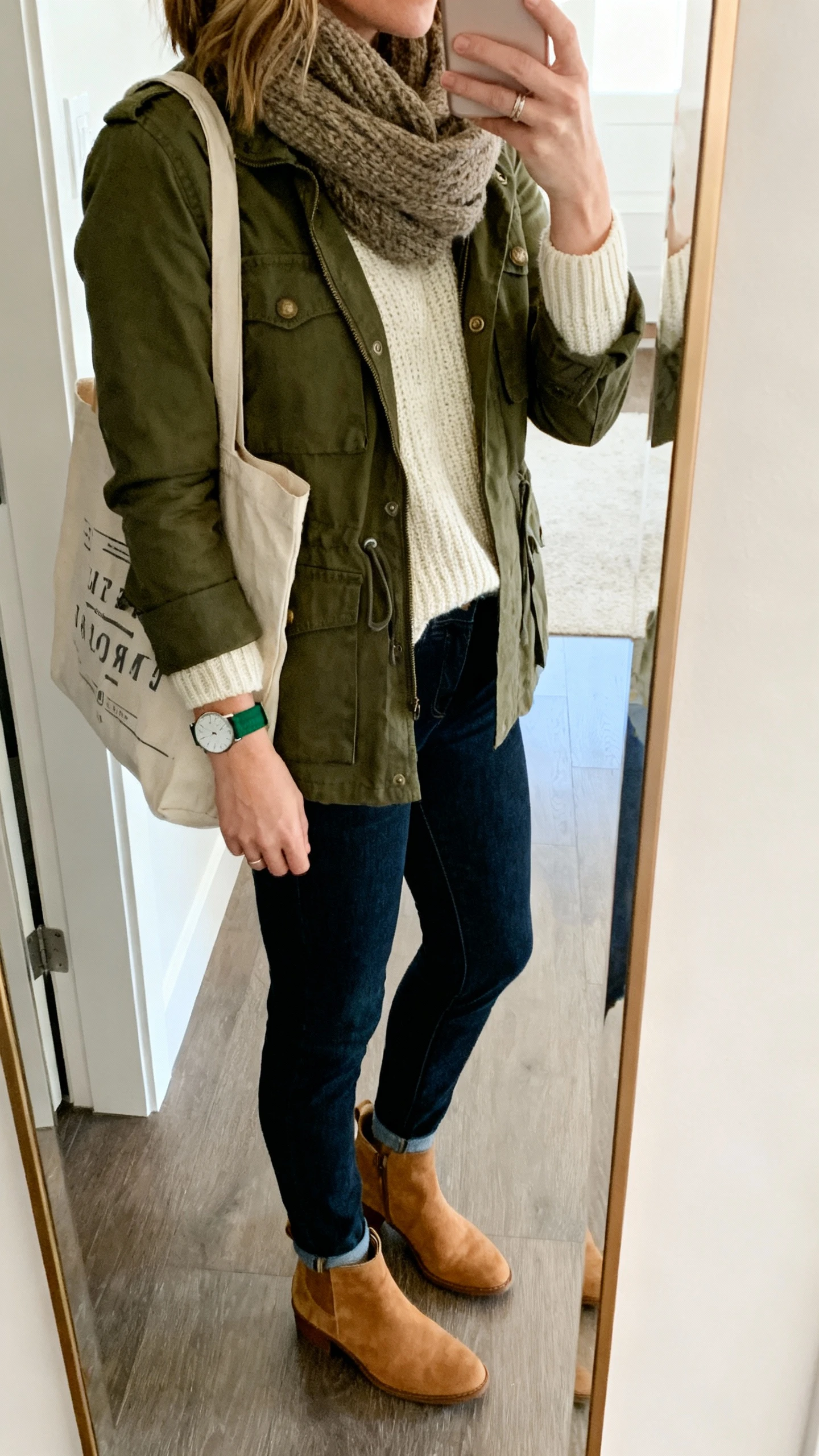 iPhone mirror selfie of a woman in a smart-casual layered look: olive utility jacket, cream knit sweater, dark indigo straight jeans, tan suede ankle boots, minimalist field-style watch with green fabric strap, wool scarf, canvas tote, face not visible, side angle, entryway full-length mirror, natural daylight, iPhone photo quality.