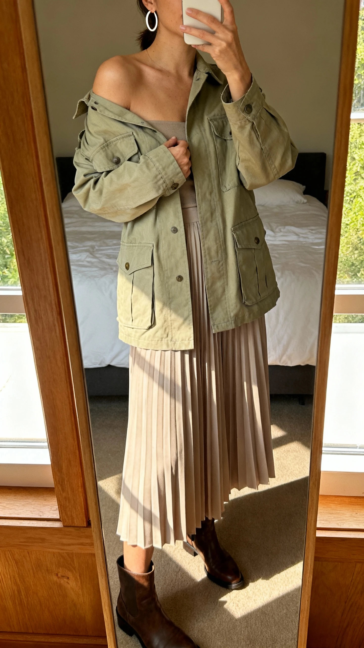 iPhone mirror selfie of a woman in a soft utility look: field jacket layered over a pleated midi skirt, ankle boots, simple hoop earrings, face not visible, cropped at shoulders, bedroom mirror, warm afternoon window light, iPhone photo quality.
