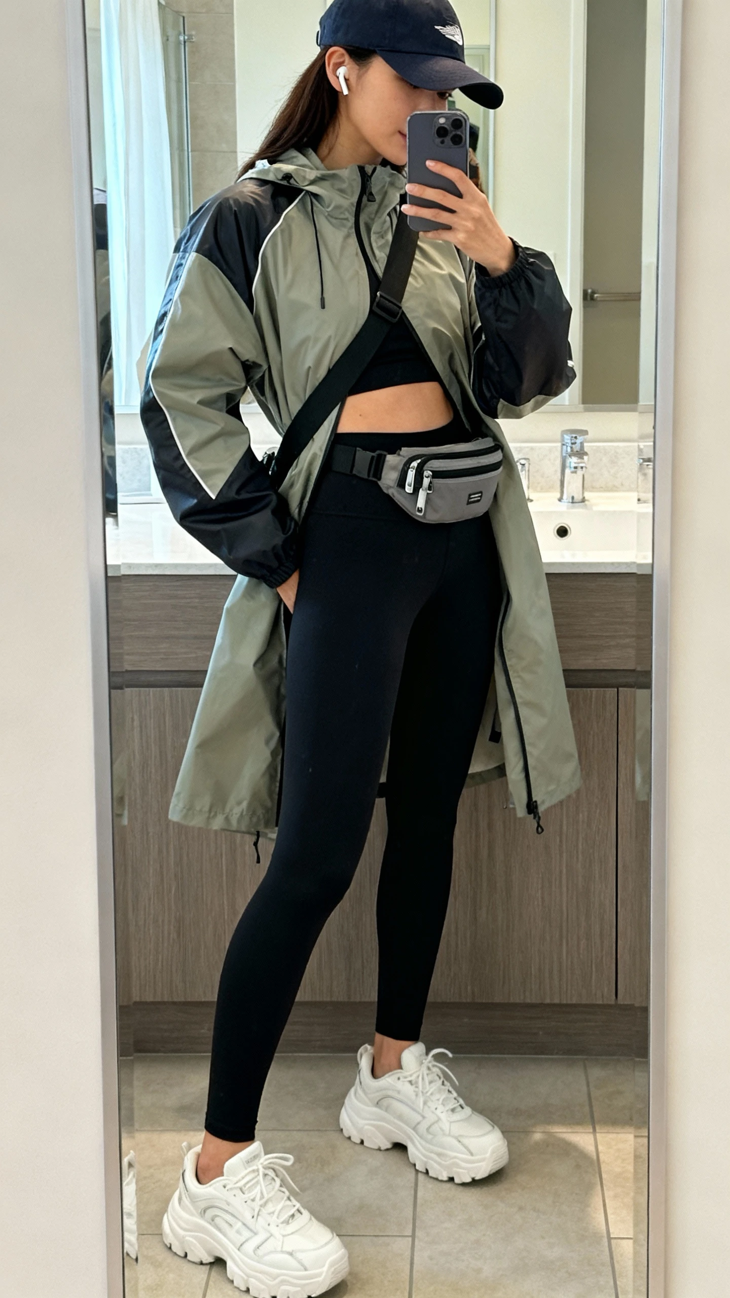iPhone mirror selfie of a woman in a sporty-chic raincoat, black high-waisted leggings, chunky white sneakers, baseball cap, crossbody belt bag, and wireless earbuds, face not visible, side angle in a bathroom full-length mirror with morning light, iPhone photo quality.