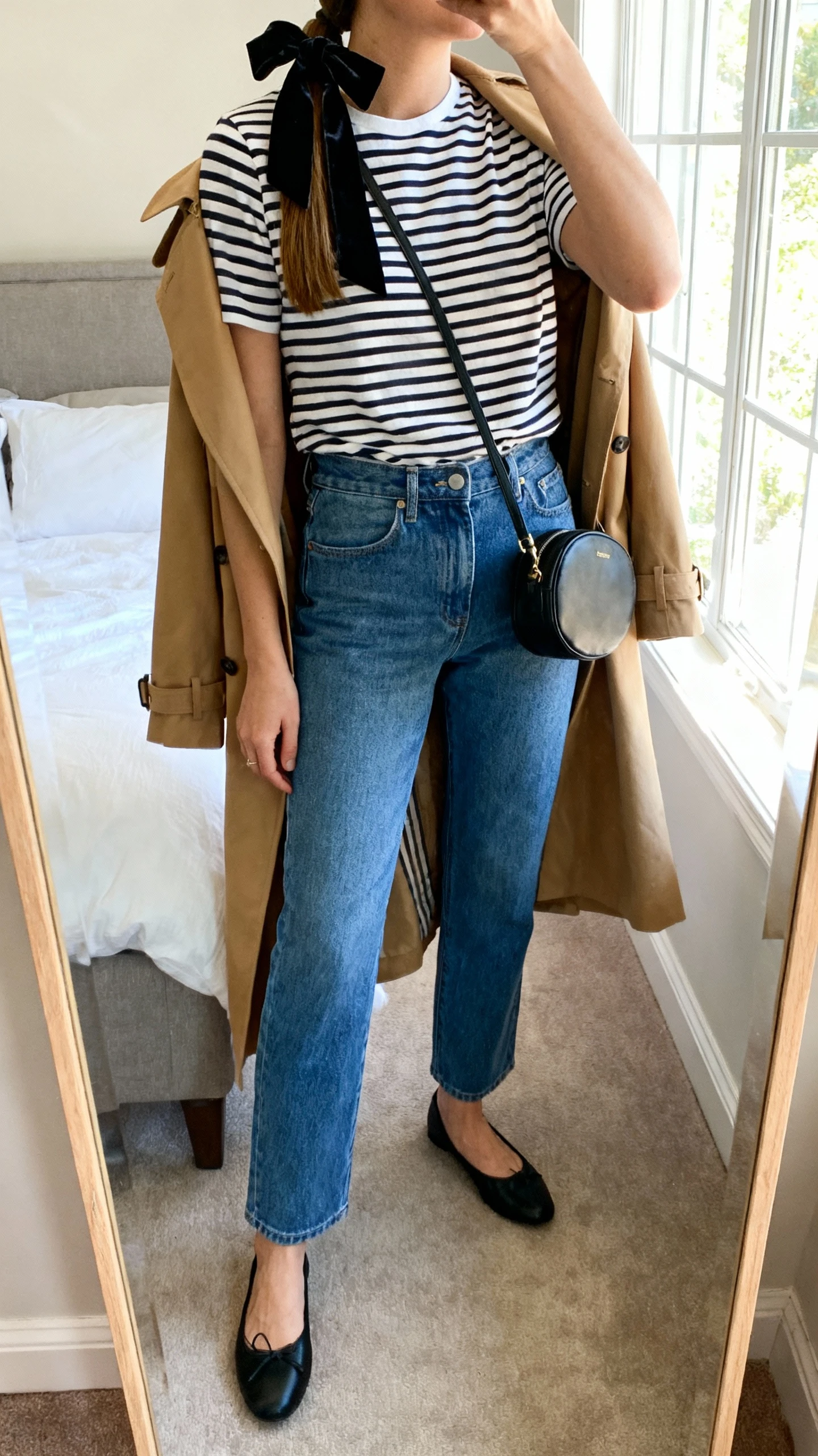 iPhone mirror selfie of a woman in a striped Breton tee, high-waisted straight-leg blue jeans, black ballet flats, camel trench coat draped over shoulders, small black crossbody bag, and a black velvet ribbon bow tied in her low ponytail; face not visible, cropped at neck, bedroom mirror, morning window light, iPhone photo quality.