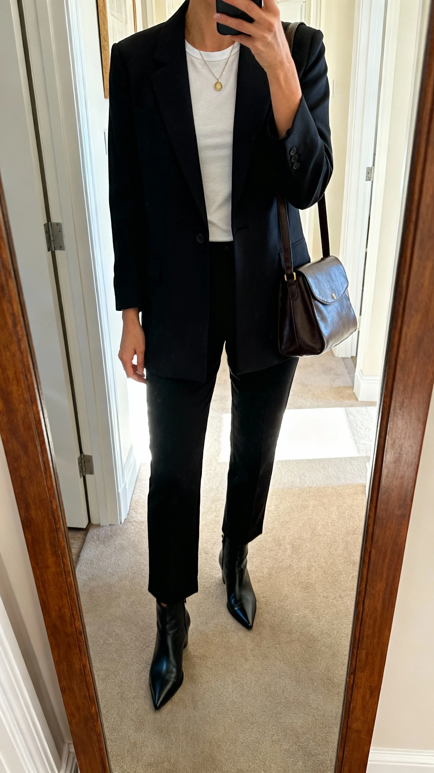 iPhone mirror selfie of a woman in a tailored blazer over a fitted tee, straight black trousers, pointed ankle boots, subtle pendant necklace, carrying a medium leather shoulder bag that works for office to weekend; face not visible, cropped at shoulders, entryway full-length mirror, bright natural light, iPhone photo quality.
