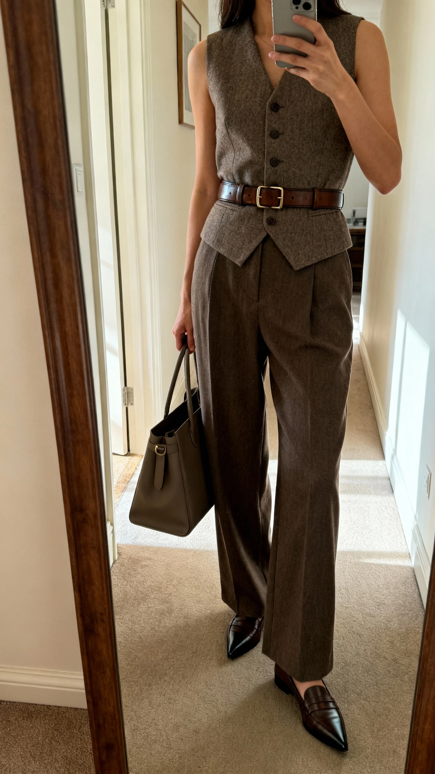 iPhone mirror selfie of a woman in a tailored vest and wide-leg trousers, sleek leather belt, pointed-toe loafers, structured tote in hand, face not visible, side angle in a hallway full-length mirror with indoor daylight, iPhone photo quality.