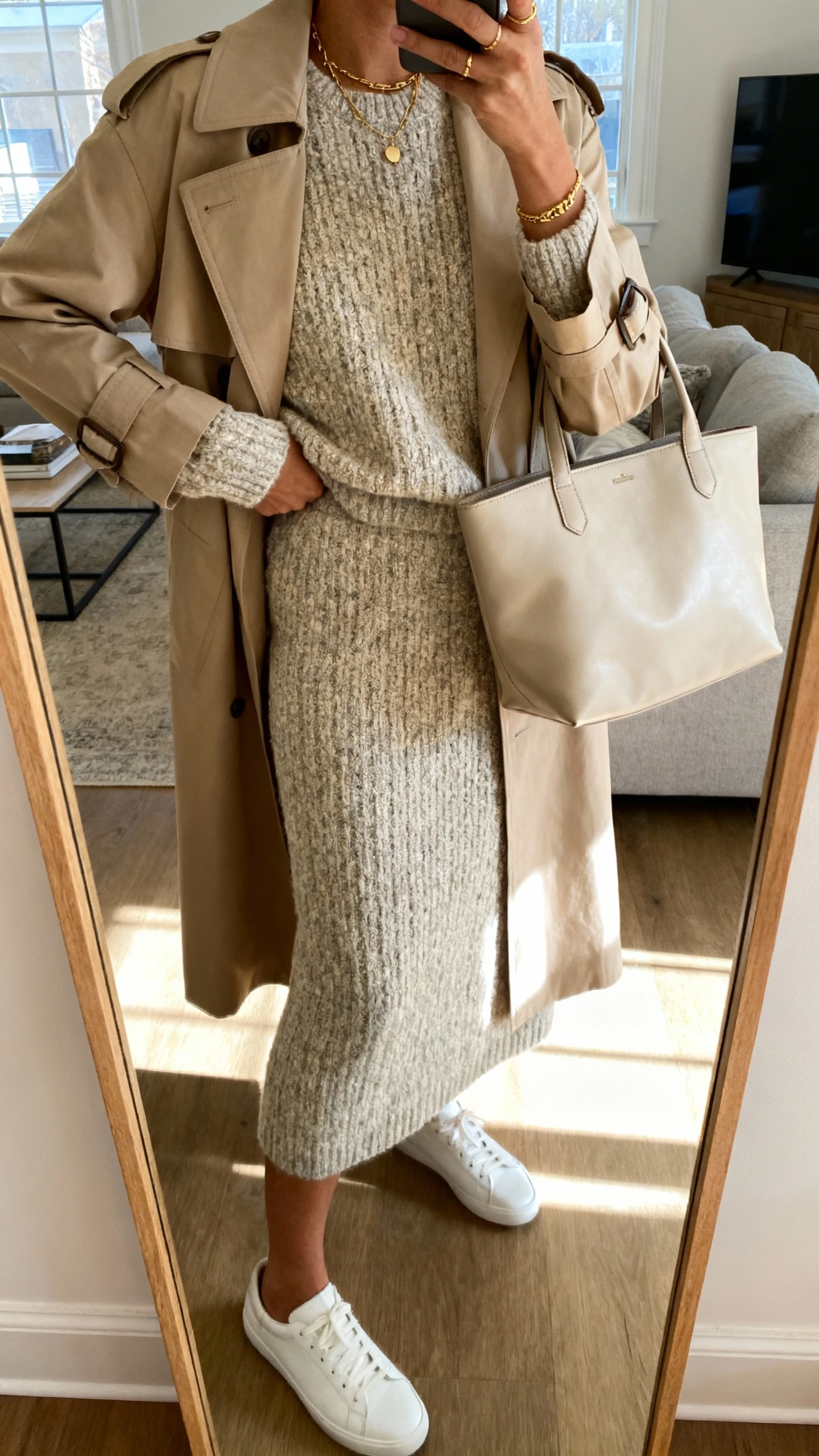 iPhone mirror selfie of a woman in a textured knit set (matching sweater and skirt) with a beige trench, minimalist white sneakers, delicate gold jewelry, and a soft leather tote, face not visible, cropped at shoulders, living-room full-length mirror with natural light, iPhone photo quality.