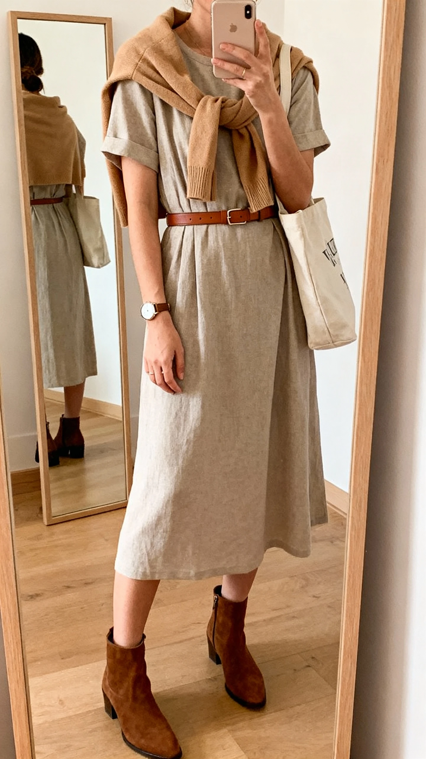 iPhone mirror selfie of a woman in a versatile capsule dress (midi, short-sleeve) layered with a camel cardigan tied over shoulders, classic ankle boots, slim leather belt, tote bag, watch, face not visible, cropped at shoulders, full-length mirror, warm indoor lighting, iPhone photo quality.