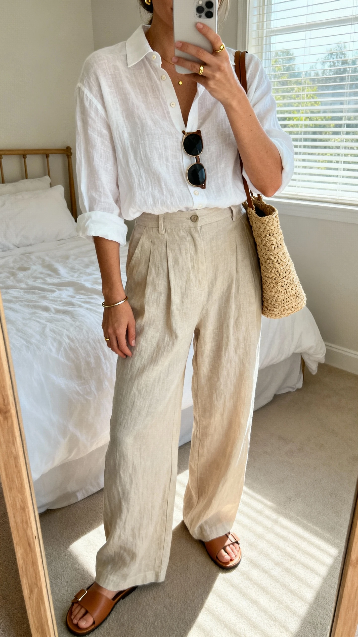 iPhone mirror selfie of a woman in a white linen button-down tucked into wide-leg linen trousers, minimalist leather slides, a raffia tote, simple gold studs, a thin bangle, and classic sunglasses; face not visible, cropped at shoulders, bedroom mirror with natural window light, iPhone photo quality.