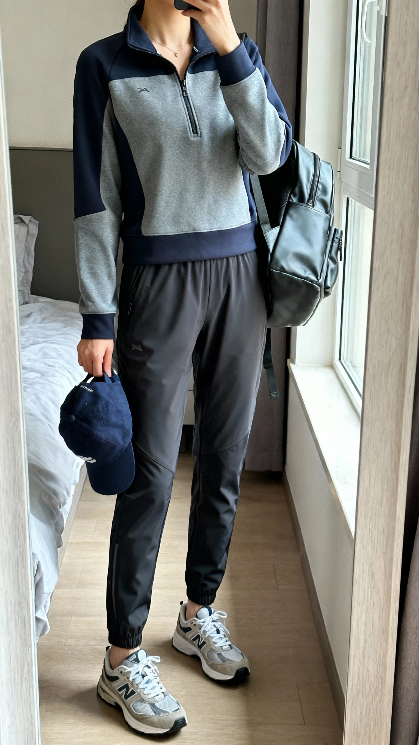 iPhone mirror selfie of a woman in an athleisure set: sporty half-zip pullover, tapered tech pants, retro trainers, baseball cap tucked in hand, and a sleek backpack; face not visible, cropped at neck, bedroom mirror with natural window light, iPhone photo quality.