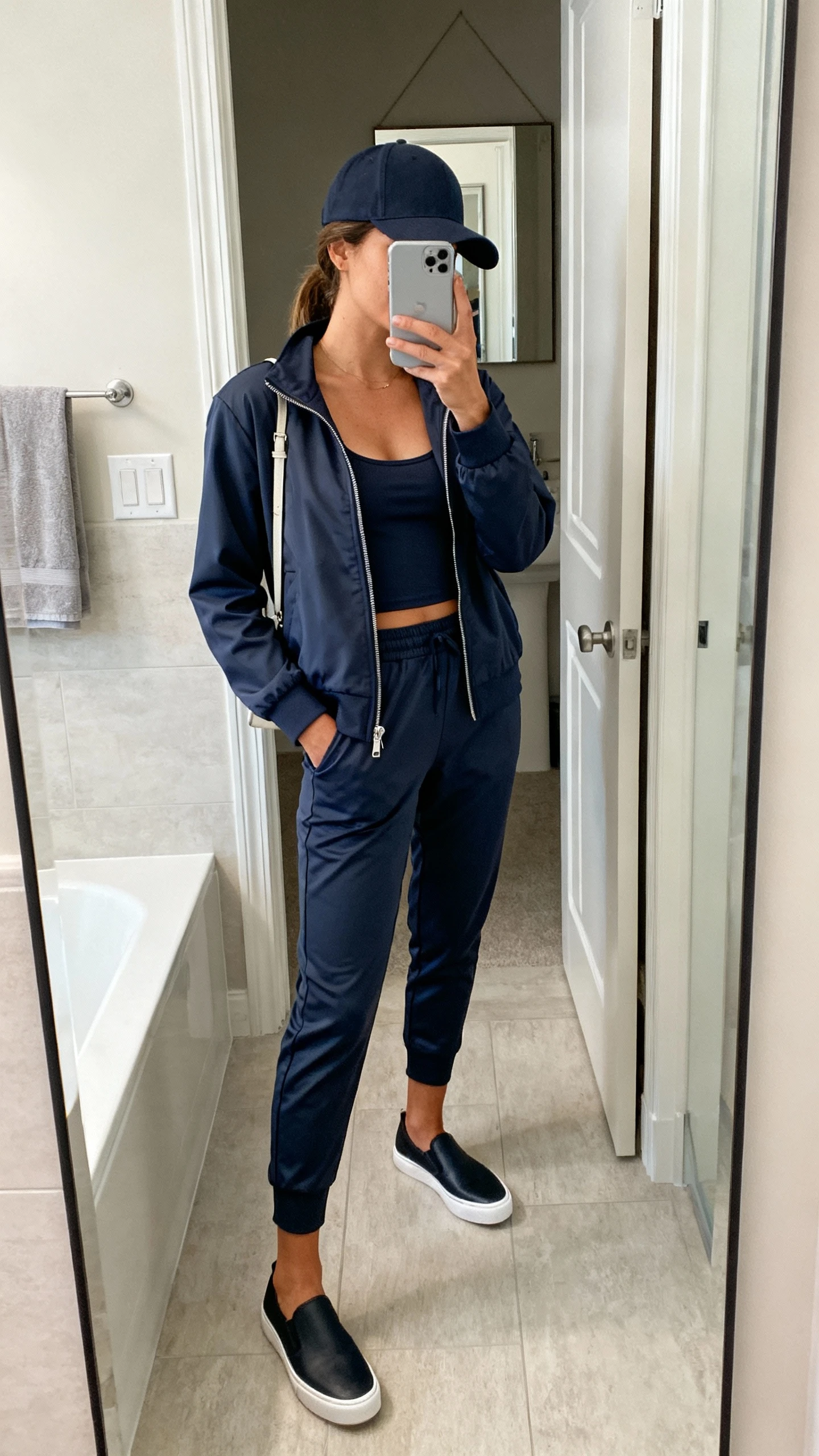 iPhone mirror selfie of a woman in an elevated athleisure uniform: wrinkle-resistant matching zip-up track jacket and tapered joggers, breathable tank underneath, sleek slip-on trainers, structured baseball cap, minimal crossbody, face not visible, bathroom full-length mirror, indoor soft lighting, iPhone photo quality.