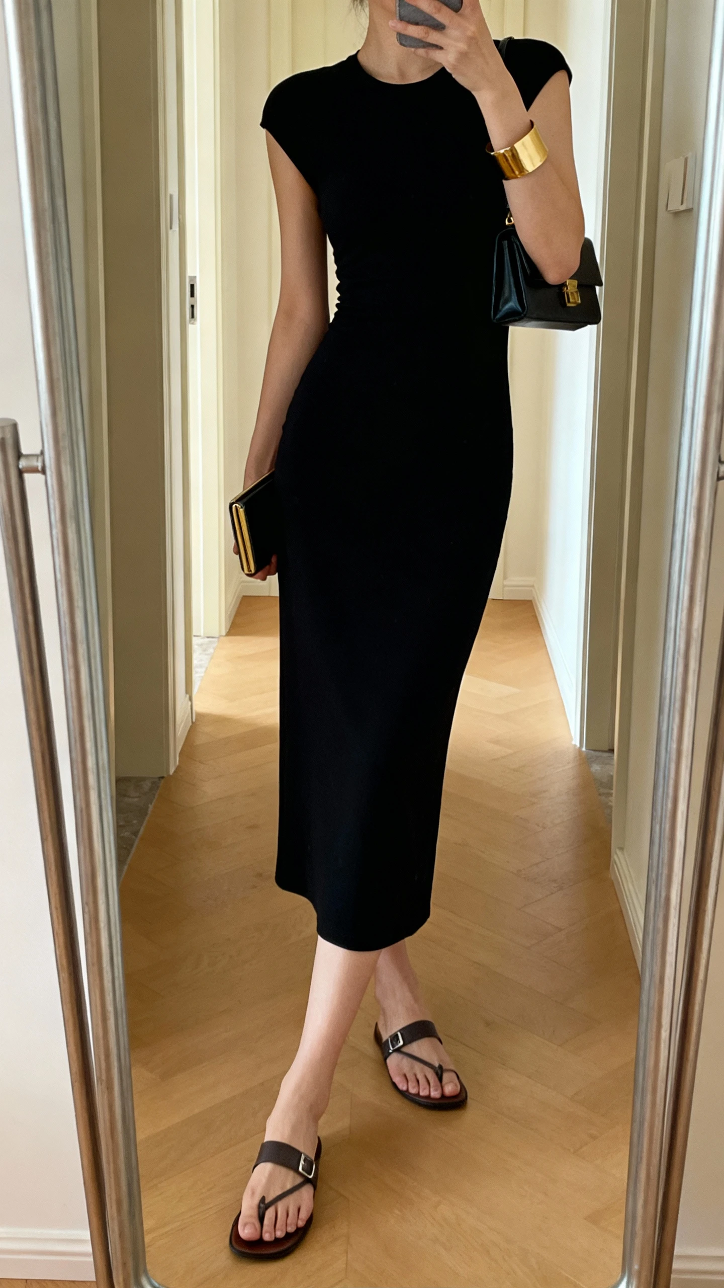 iPhone mirror selfie of a woman in an elevated black dress (midi, streamlined), walkable flat sandals, small clutch or mini shoulder bag, gold cuff bracelet, face not visible, cropped at neck, entryway full-length mirror with warm indoor lighting, iPhone photo quality