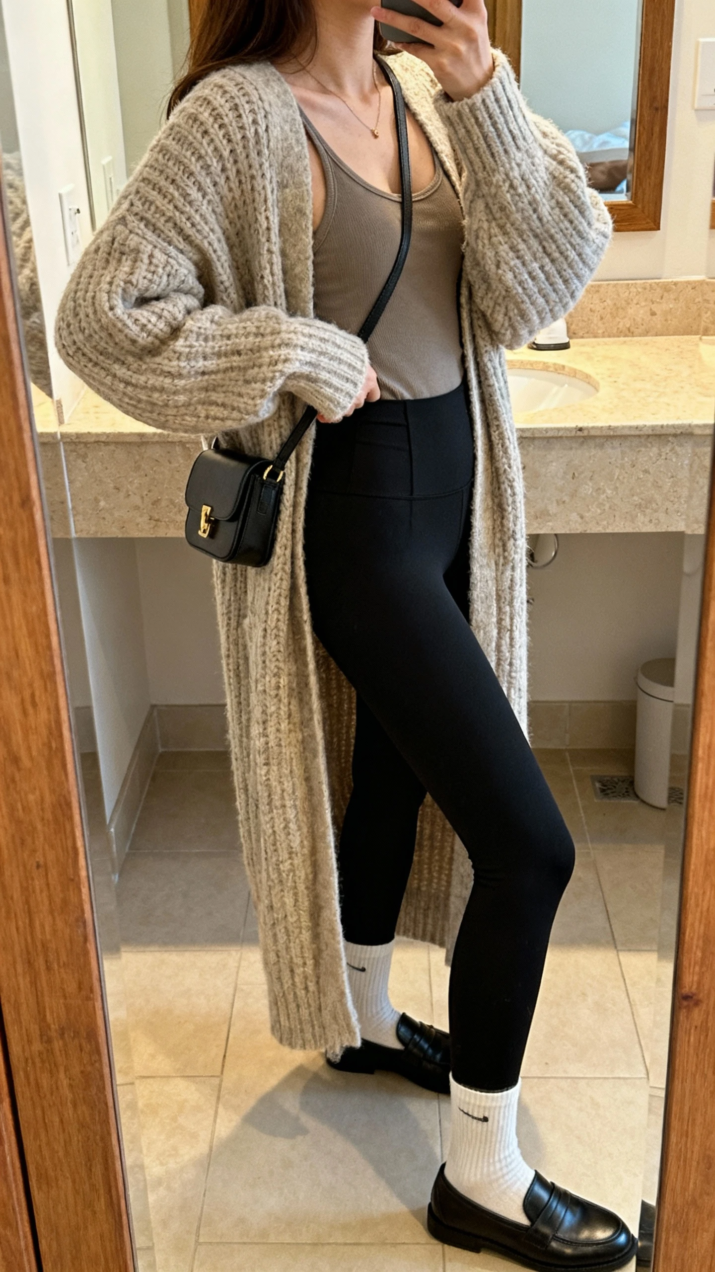 iPhone mirror selfie of a woman in an oversized chunky cardigan layered over a longline tank, black high-waisted leggings, ankle socks, and slip-on loafers, small crossbody bag on shoulder, face not visible, side angle in bathroom mirror, warm indoor lighting, iPhone photo quality