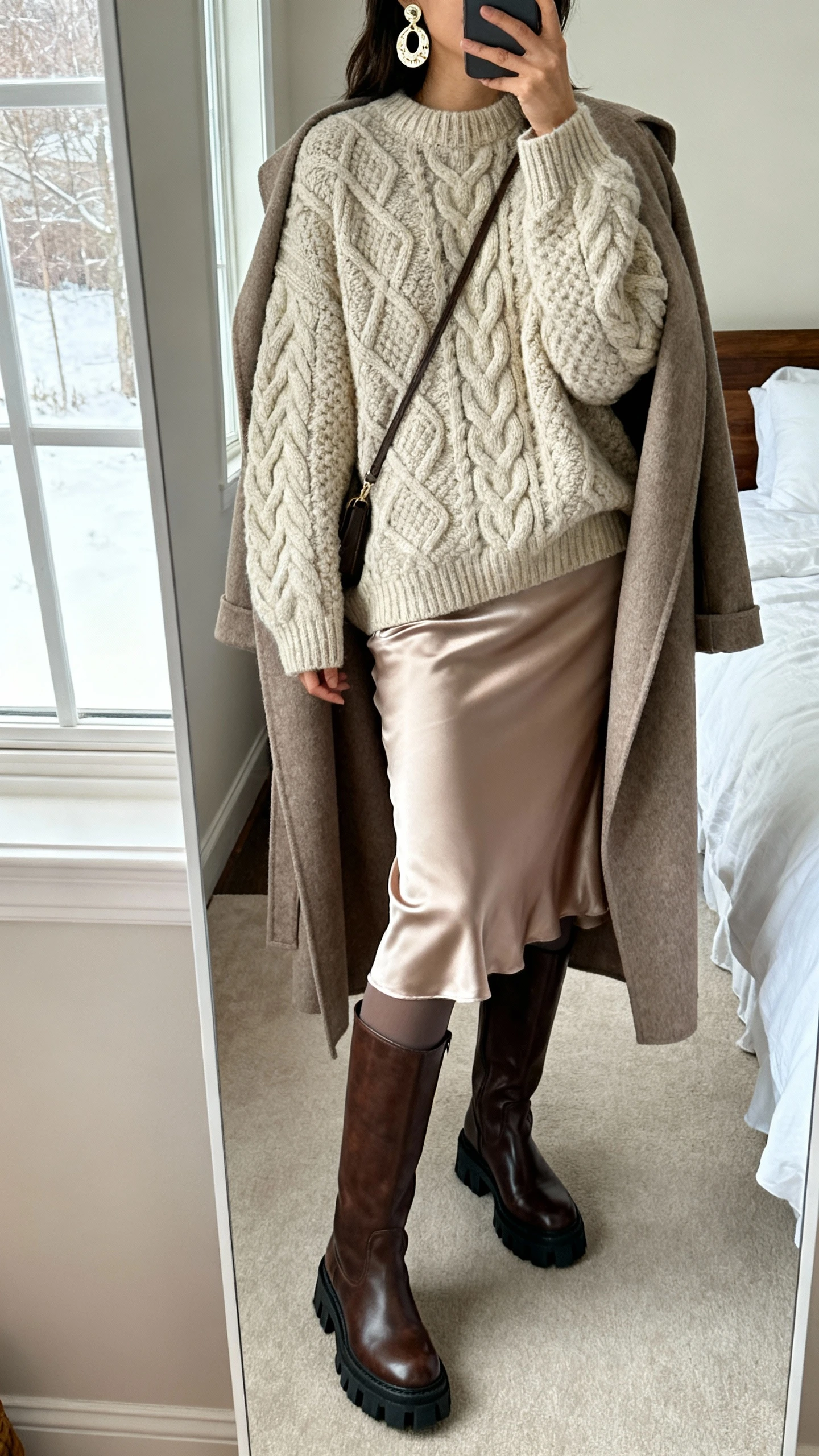 iPhone mirror selfie of a woman in an oversized chunky cable knit sweater, bias-cut satin midi slip skirt, opaque tights, leather knee-high boots with sturdy sole, long wool wrap coat draped over shoulders, statement earrings and a simple crossbody, face not visible, cropped at shoulders, bedroom mirror with natural window light, iPhone photo quality.