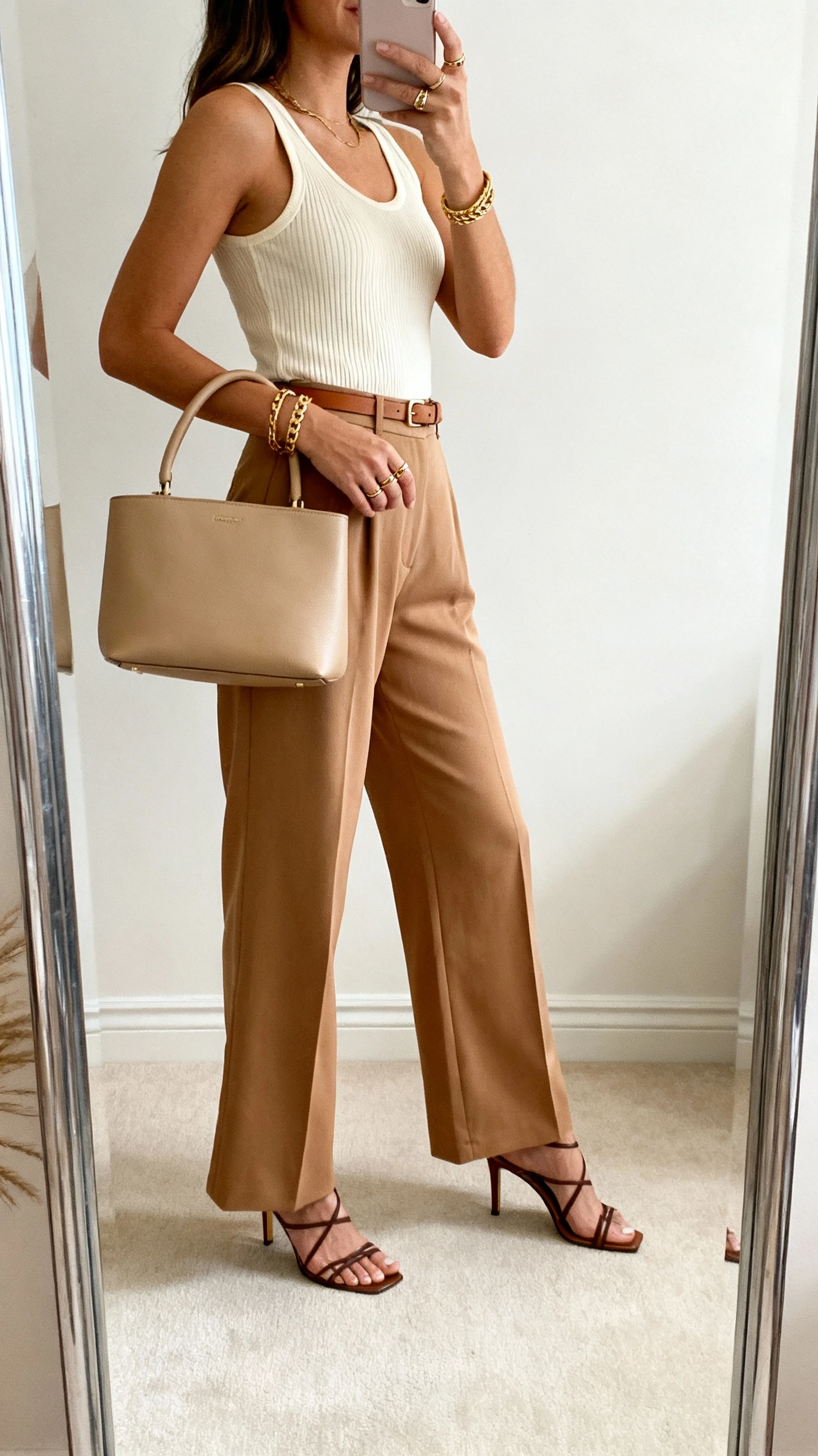 iPhone mirror selfie of a woman in camel wide-leg trousers with a fitted cream ribbed tank, thin leather belt, heeled strappy sandals, structured beige top-handle bag, layered gold bracelet and ring stack, face not visible, side angle in full-length mirror, soft indoor lighting, iPhone photo quality.