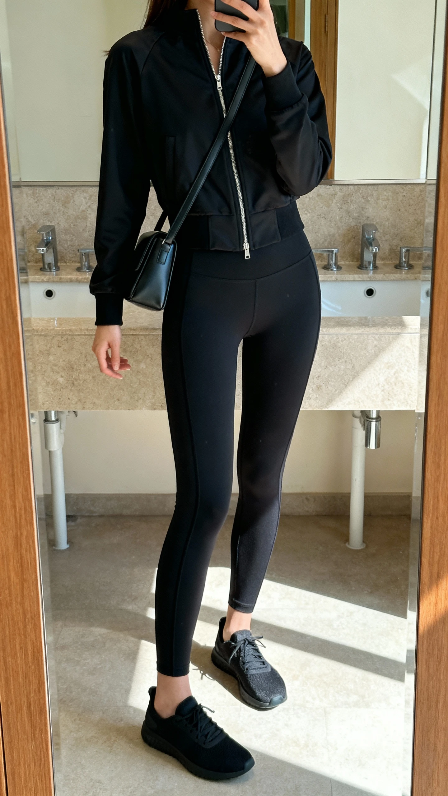 iPhone mirror selfie of a woman in elevated athleisure: black streamlined leggings, a sleek black zip-up jacket, minimalist sneakers, and a slim sleek shoulder bag, face not visible, bathroom mirror, indoor daylight, iPhone photo quality.