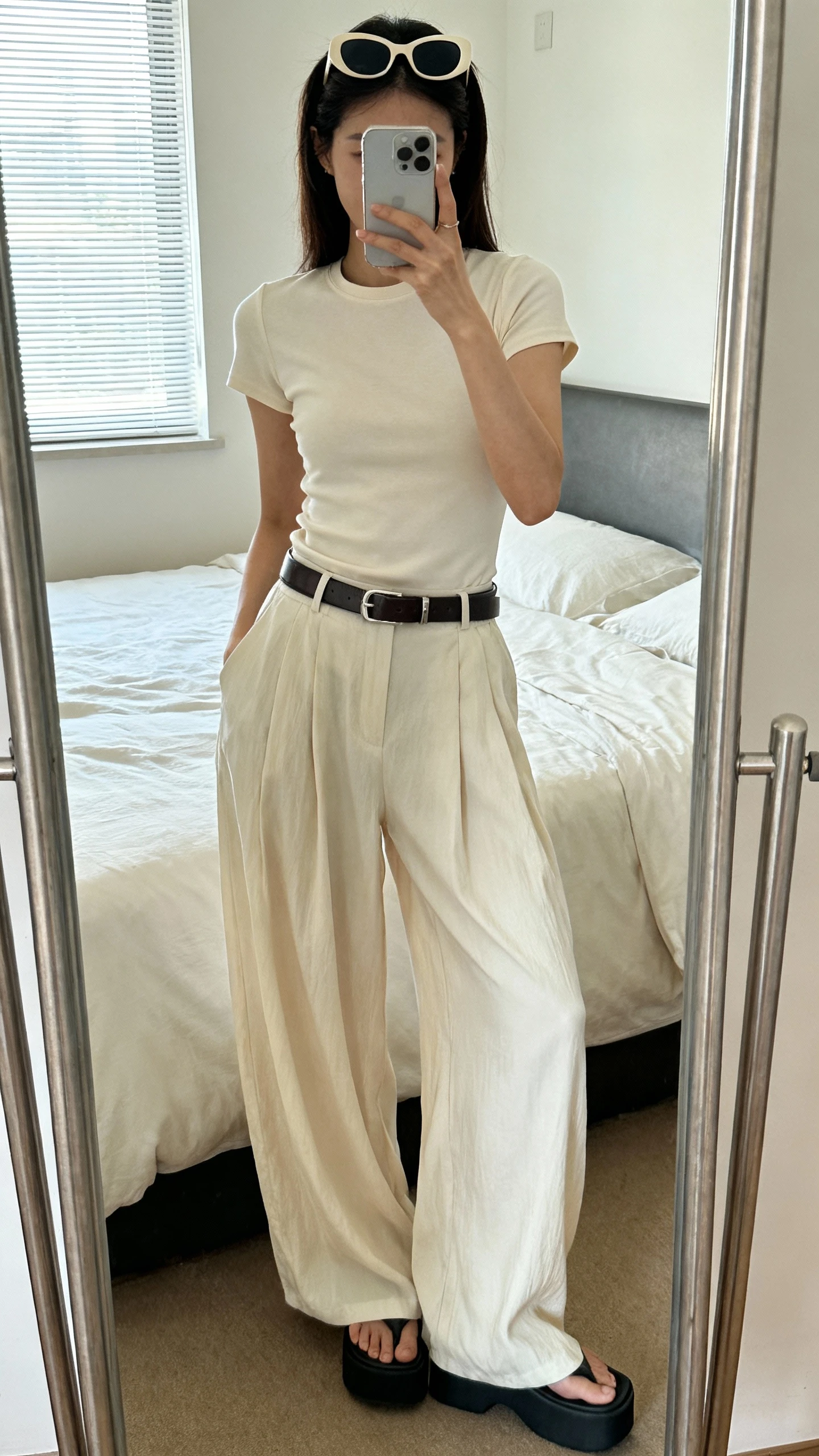 iPhone mirror selfie of a woman in flowy wide-leg pants and a fitted baby tee, sleek belt, retro sunglasses pushed up on head, platform sandals, face not visible, cropped at shoulders, bedroom mirror with natural window light, iPhone photo quality