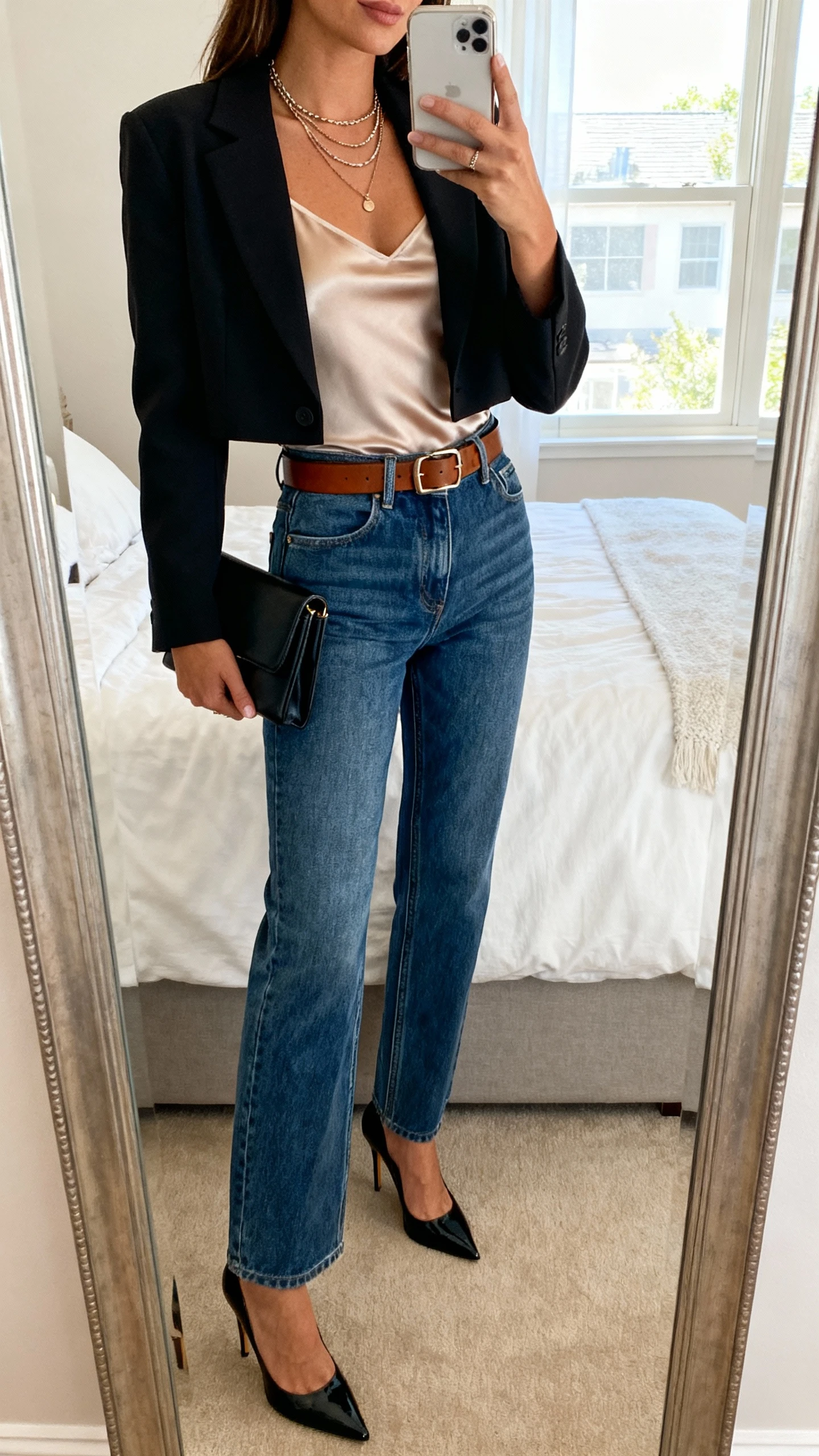iPhone mirror selfie of a woman in high-rise straight-leg jeans, a fitted bodysuit or silky cami, sleek heels, leather belt, and a cropped blazer, with a simple clutch and layered necklaces, face not visible, cropped at shoulders, bedroom mirror, natural window light, iPhone photo quality.